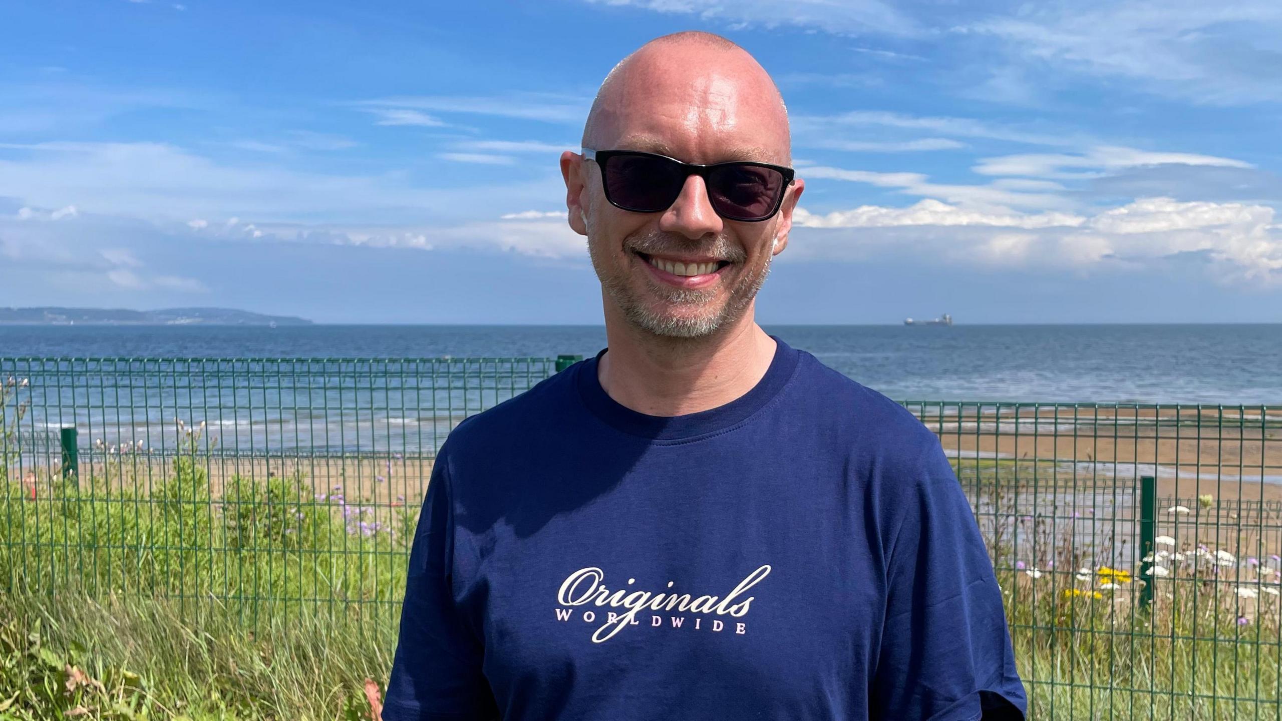 A man wearing a navy t-shirt and sunglasses. He is standing in front of a beach and is smiling at the camera. 