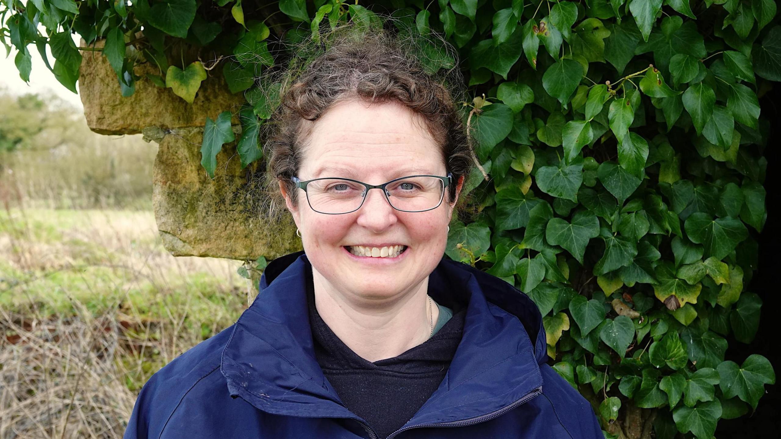 Conservationist Jenny Ashdown smiling for the camera by a wall covered with ivy. She is wearing glasses. It's an overcast day.