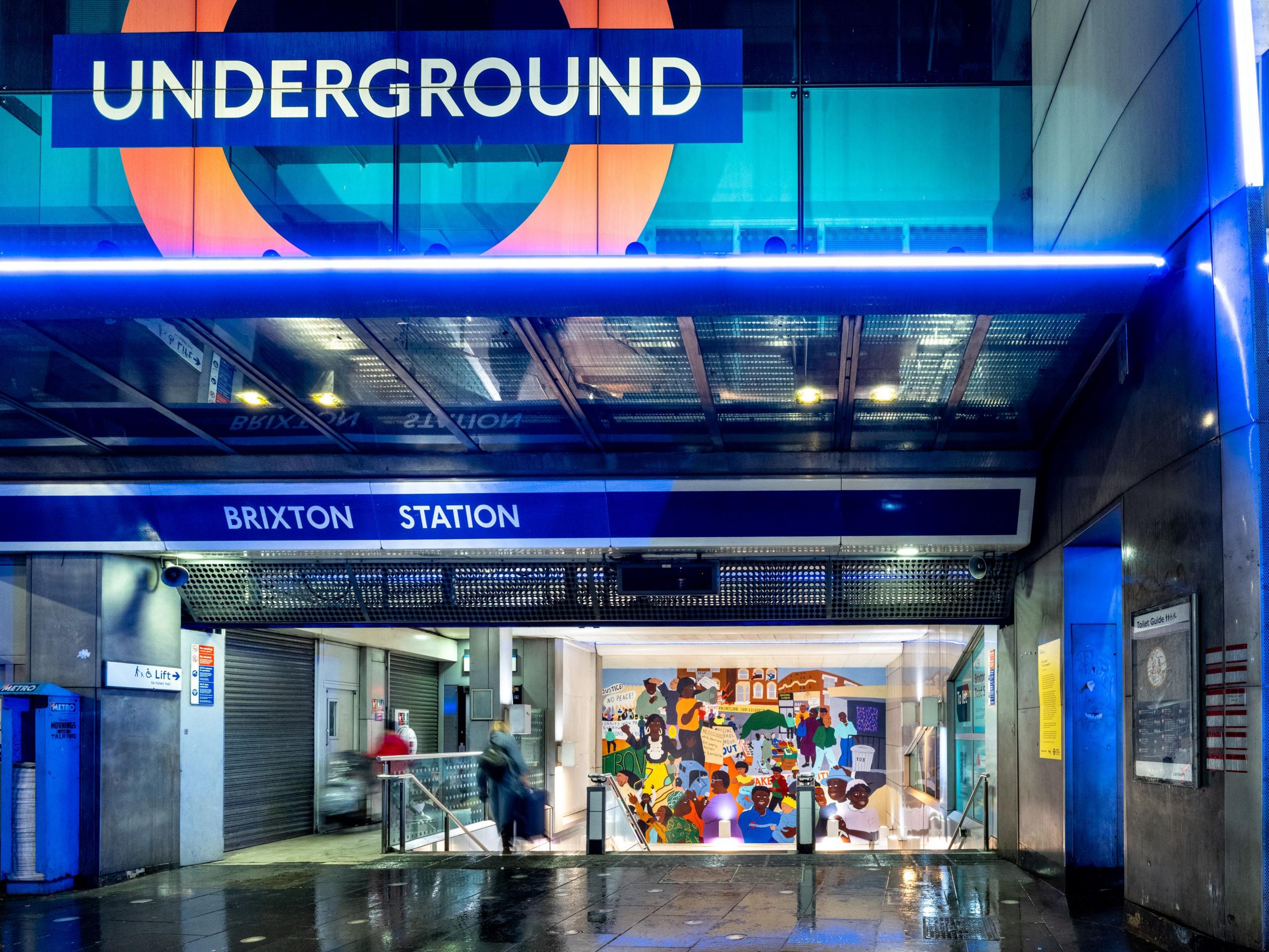 The entrance to Brixton Tube station with blue signage above the doorway. Inside the building is the vibrant mural with above a staircase leading down. The floor outside the station looks wet.