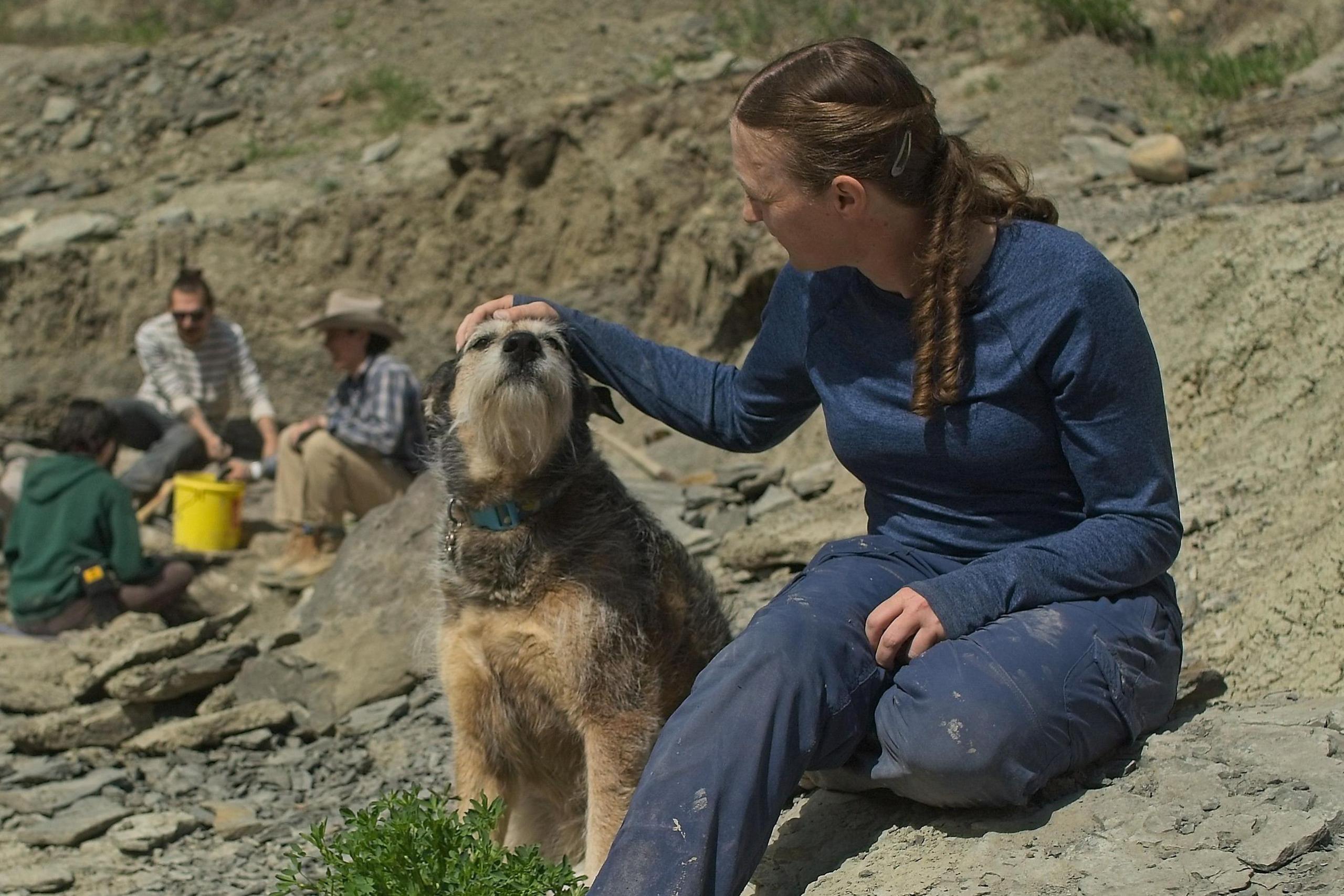 Prof Bamforth's dog, Aster, sits next to her on rocks as she strokes the dog's head, with people digging for fossils in the background.
