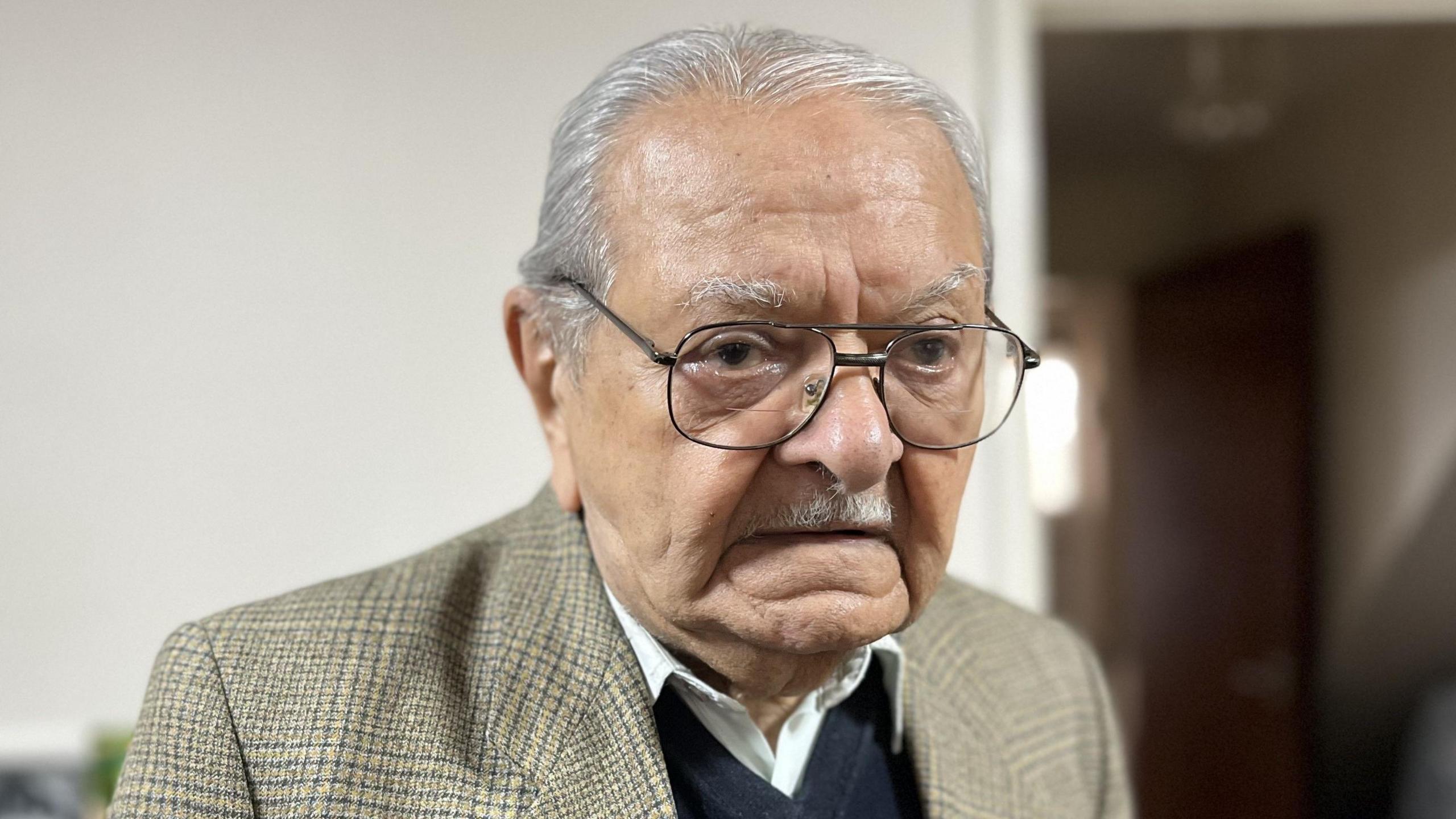 A portrait image of Mr Osman. He is 91 and wearing a brown/grey checked blazer with a dark blue swearer and revealing the collars of a white shirt.