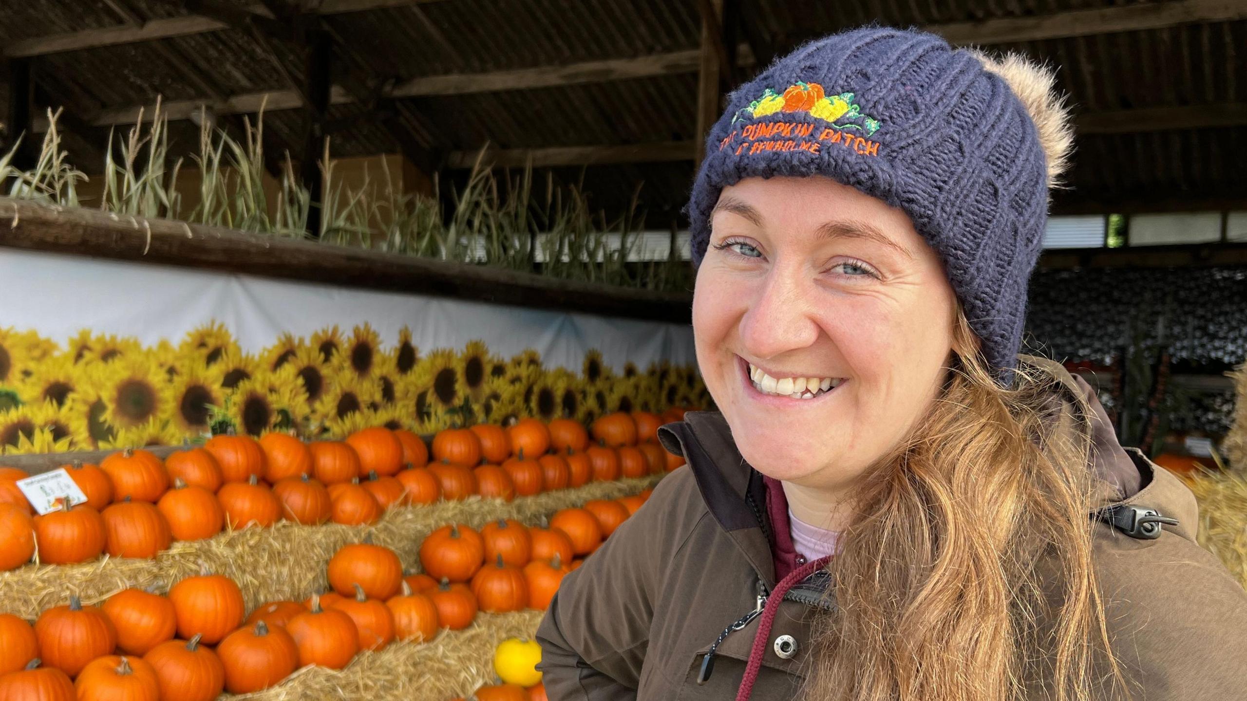 Becky Butler stands in front of rows of orange pumpkins and yellow and brown sunflowers in a barn at Pumpkin Patch Produce, Bewholme. She is wearing a blue woolly hat, a brown coat and is smiling to the camera. 