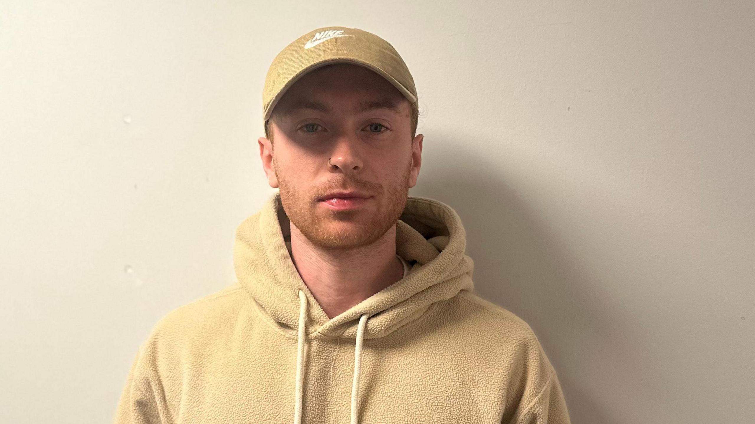 Mark is wearing a beige cap and hoodie. He has ginger facial hair and blue eyes with a silver nose piercing.