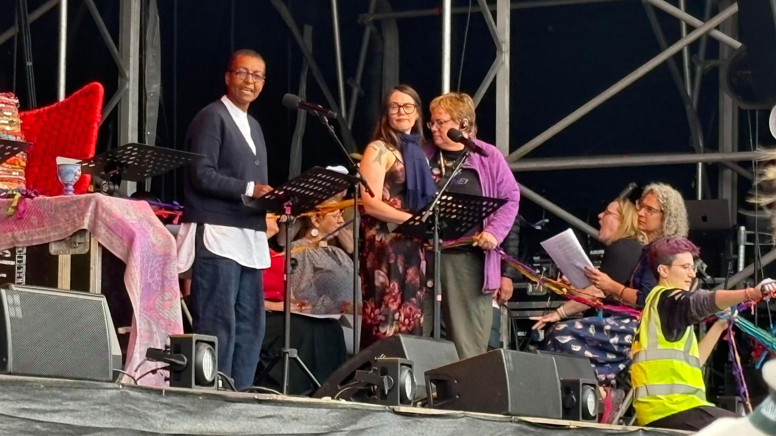 Adjoa Andoh with short dark hair and glasses, wearing a black sweater over a white top and blue trousers. She is a standing on a festival stage. There is a music stand in front of her. There are two women standing to her left behind a microphone. Two women as sitting on the right and singing from a sheet of paper. A woman in a hi-vis jacket is sitting on the stage and pointing.