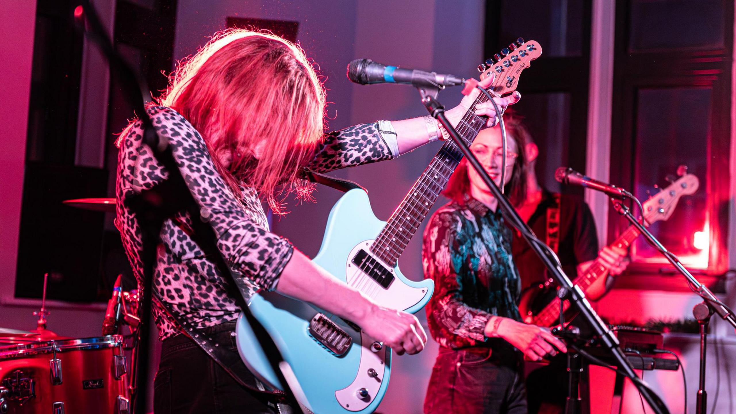 A woman with red hair plays a blue guitar. She is on stage surrounded by red lighting. She is wearing a leopard print top. Standing behind her another woman plays synth. She is wearing glasses a shirt and jeans.