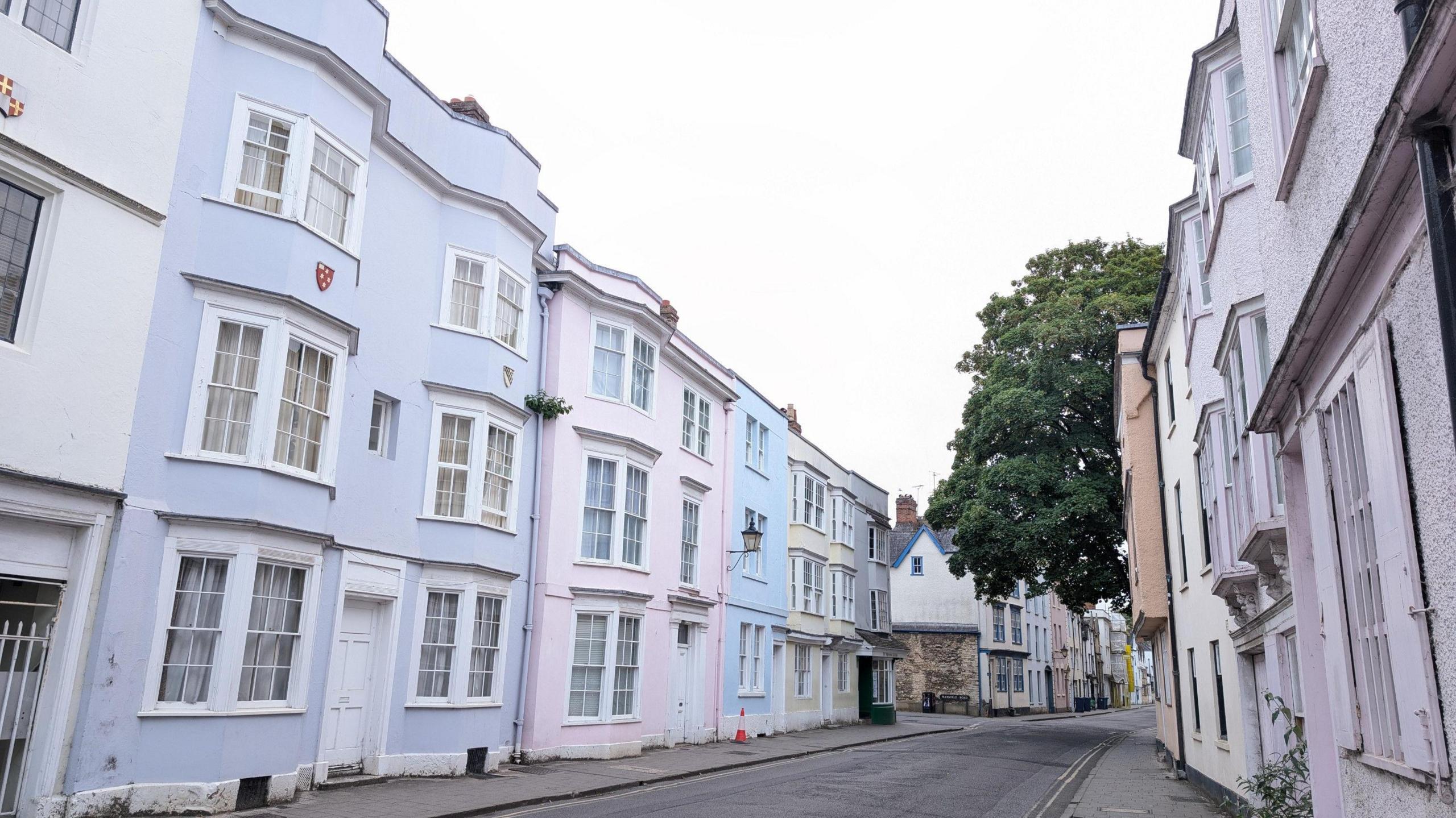 MONDAY - A street in Oxford with pastel coloured houses on each side with large traditional white windows. A large green tree overhangs the street half way down and overhead the sky is white.