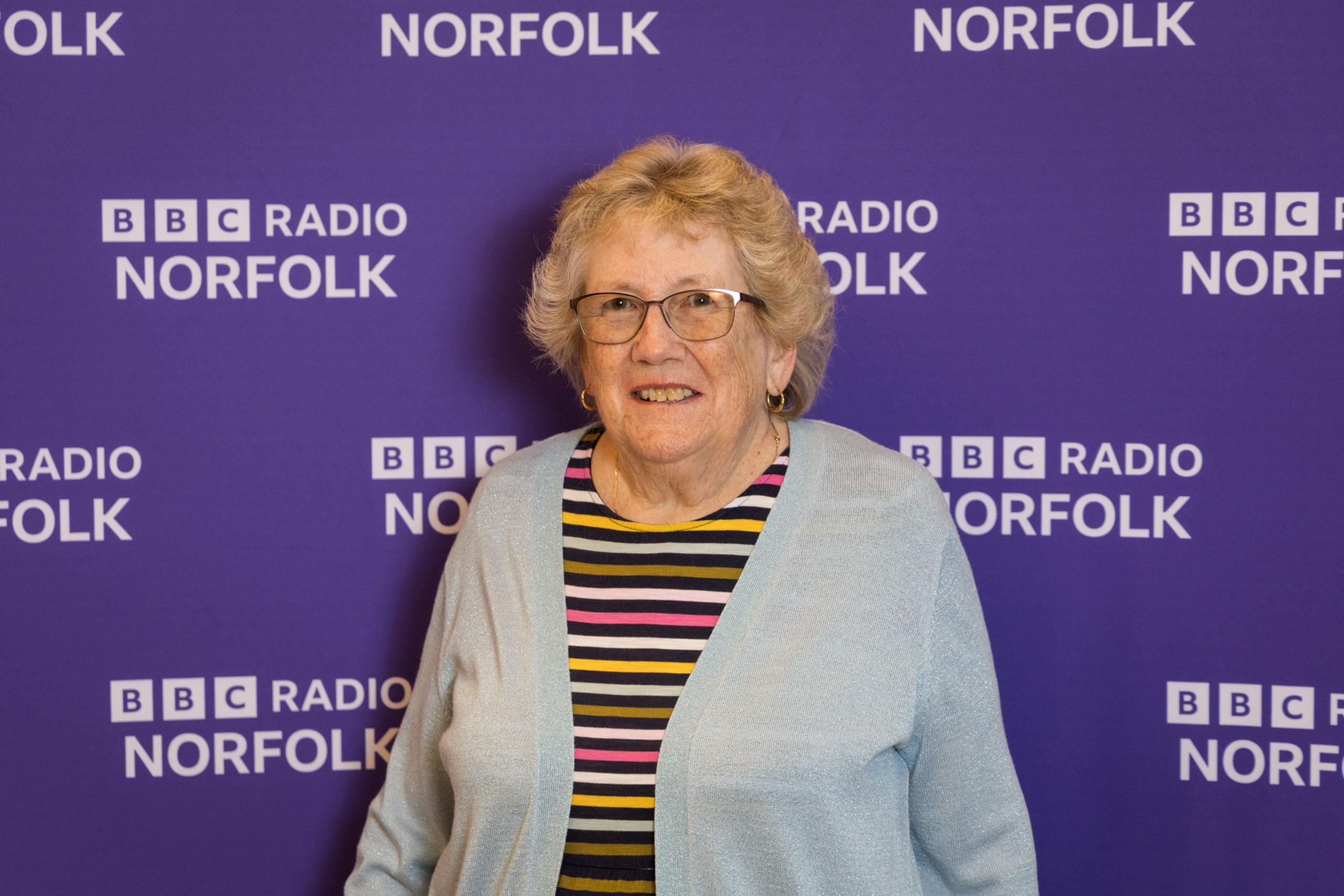 An older woman wearing a pink, yellow, white and black striped dress and a light blue cardigan stands in front of a purple background. She wears glasses and is smiling.