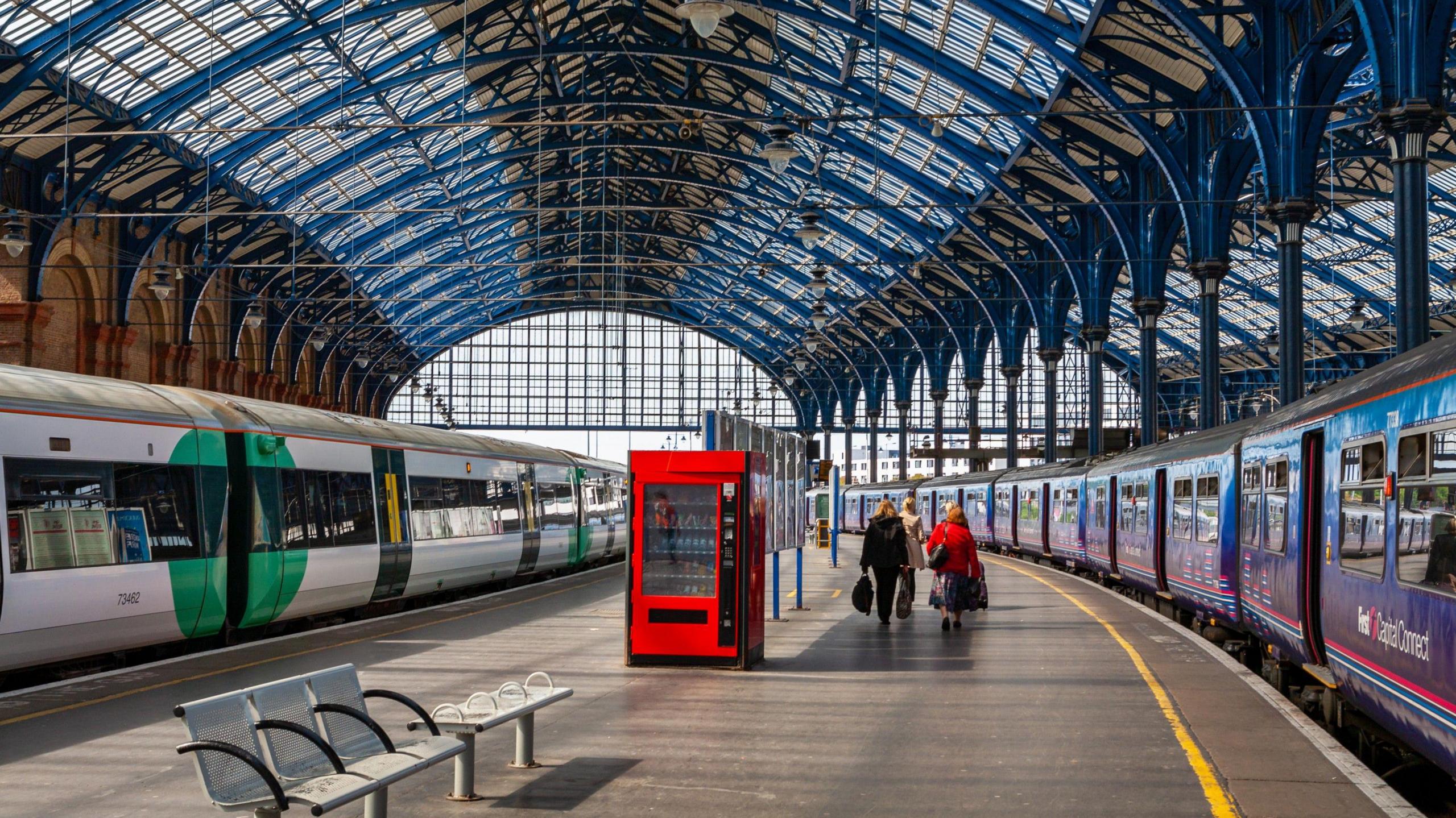 A wide view of Brighton station, with a green and white Southern Train on one side of the platform and a blue and white Capital Connect train on the other side. A group of passengers are walking away towards the turnstiles, under the large blue steel and glass canopy.