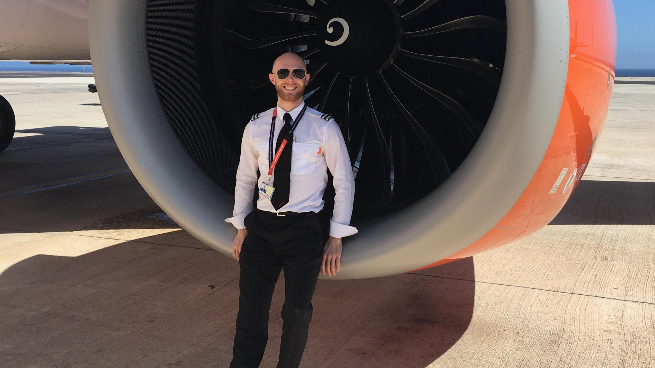 George sits at the front of one of the plane's engines wearing his pilot's uniform. He wears a white shirt and black trousers, and aviator style sunglasses. He smiles at the camera and has a brown beard and moustache.