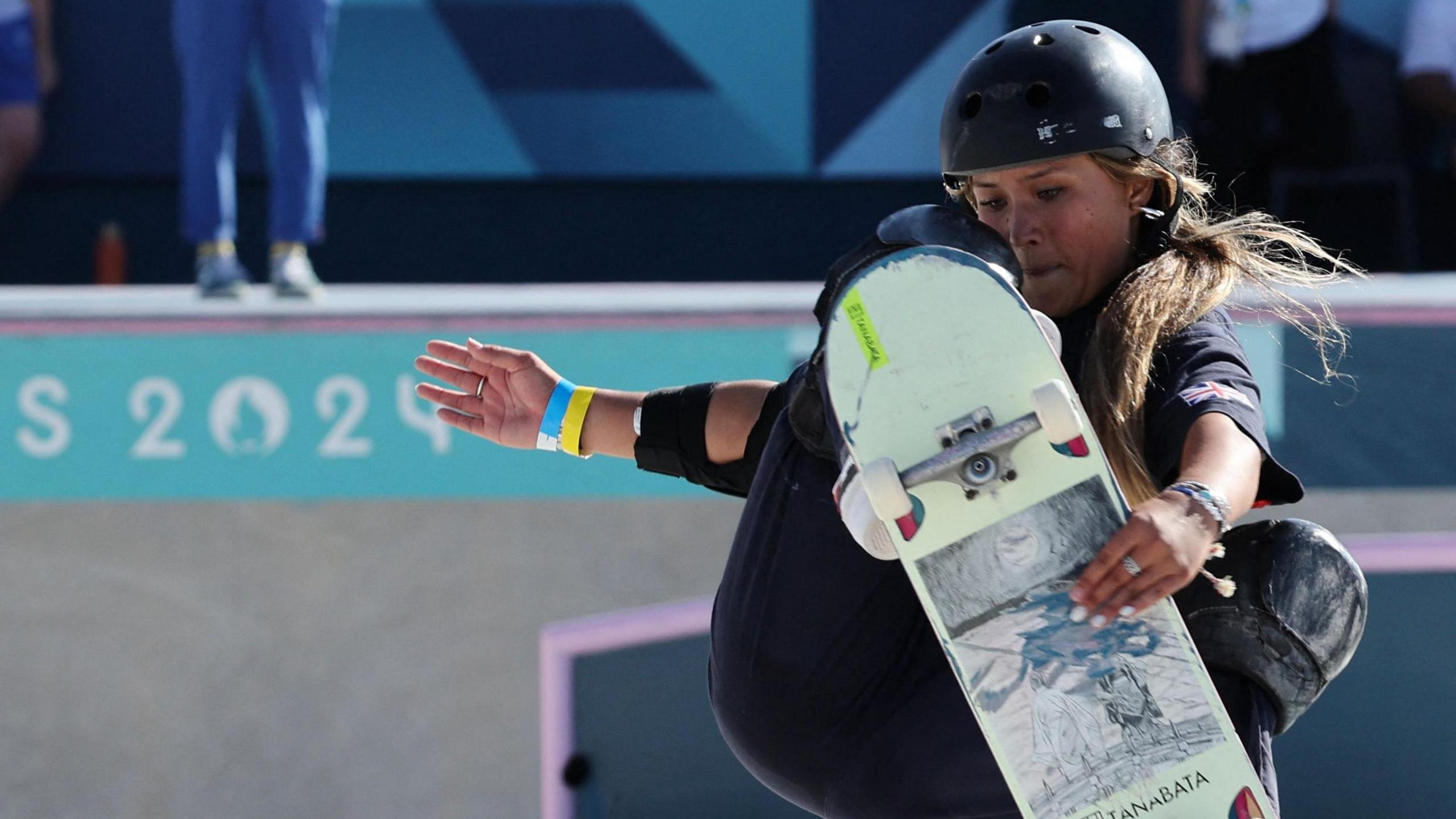 Sky Brown competing at the 2024 Olympics. She's performing a jump on her skateboard, which is mostly white underneath. She's wearing black/dark blue kit and helmet.