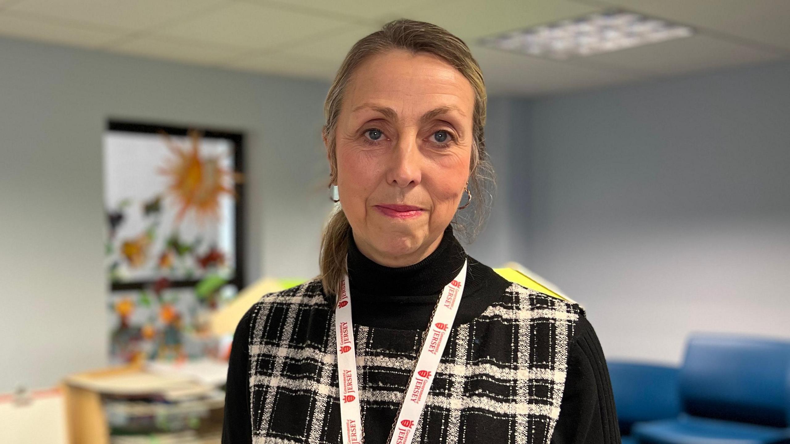 Nicola looks at the camera as she wears a black jumper with a sleeveless cardigan which has black and white crosses on in. Her hair is tied into a ponytail and behind her is children's playing equipment.