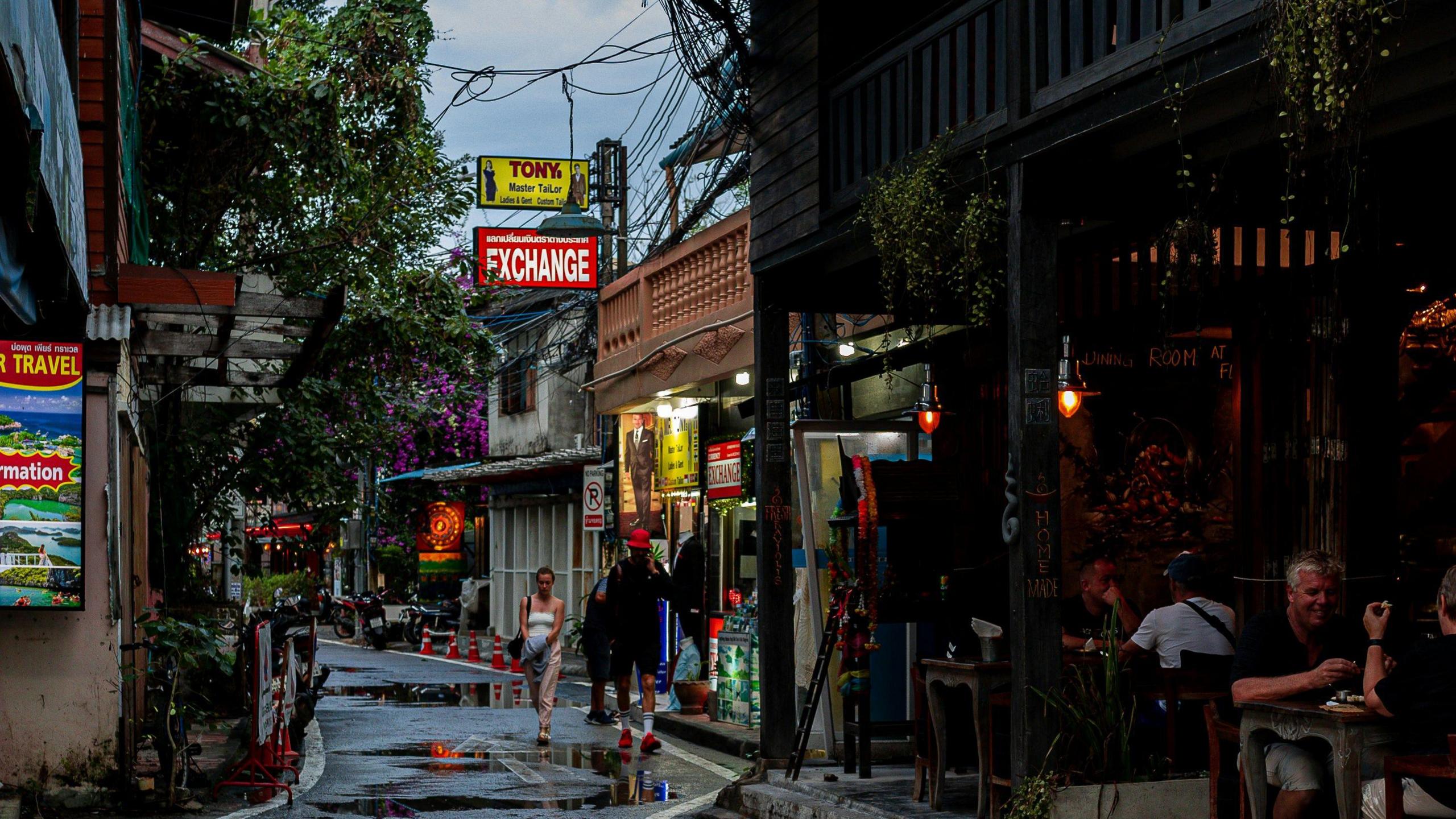 A picture of a street in Chiang Mai. There are storefronts to the right of a road adorned with various signs. One store front is open and people are sitting at tables. People are walking on the street. There are trees, foliage and cables overhead.