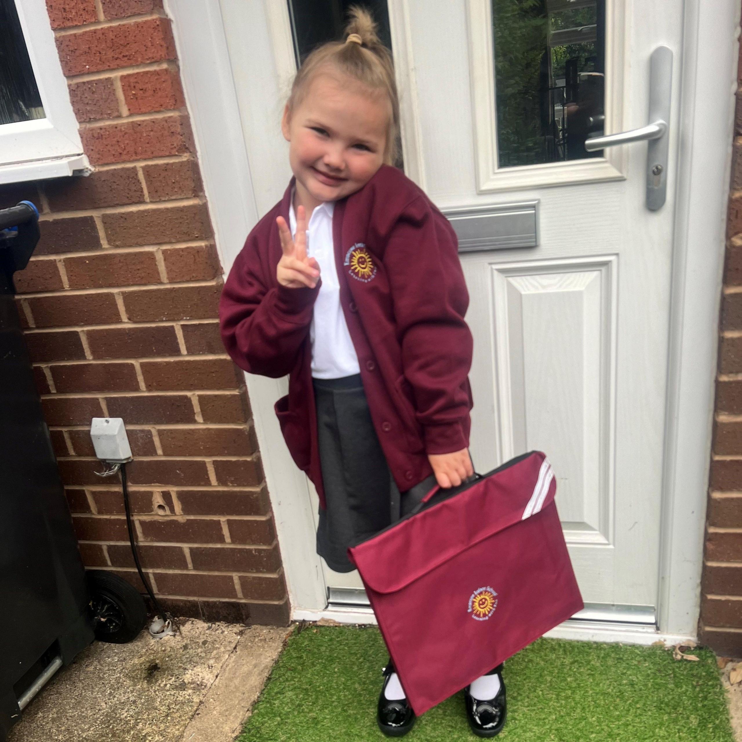 A young girl wearing a school uniform with a maroon cardigan and holding a maroon book bag.