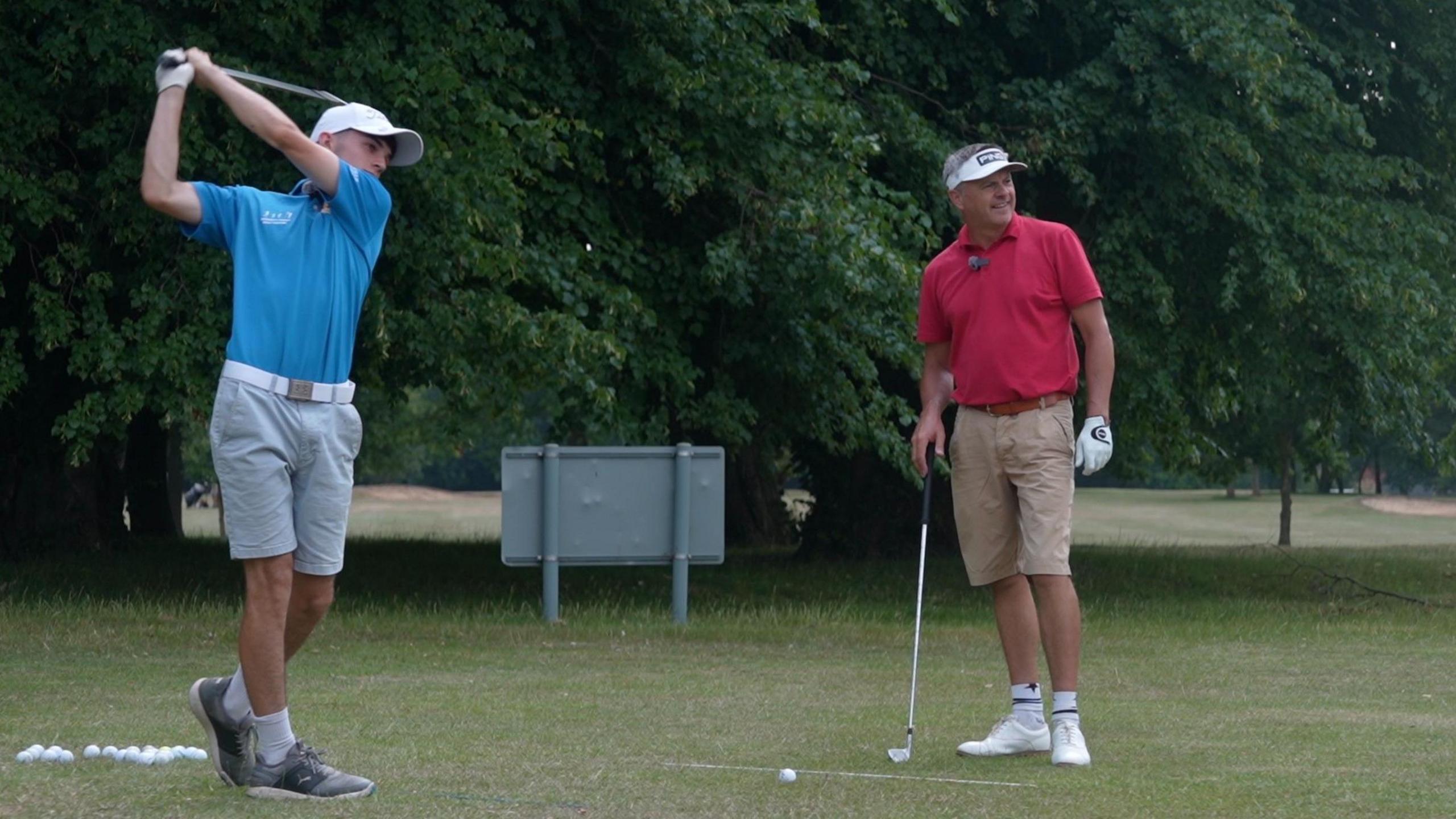 Liam, who is a left-handed golfer, hits a shot with an iron on the range at Dereham Golf Club. His club-mate Andrew smiles as he admires the shot. The pair are hitting shots off the grass with bushes and trees behind.