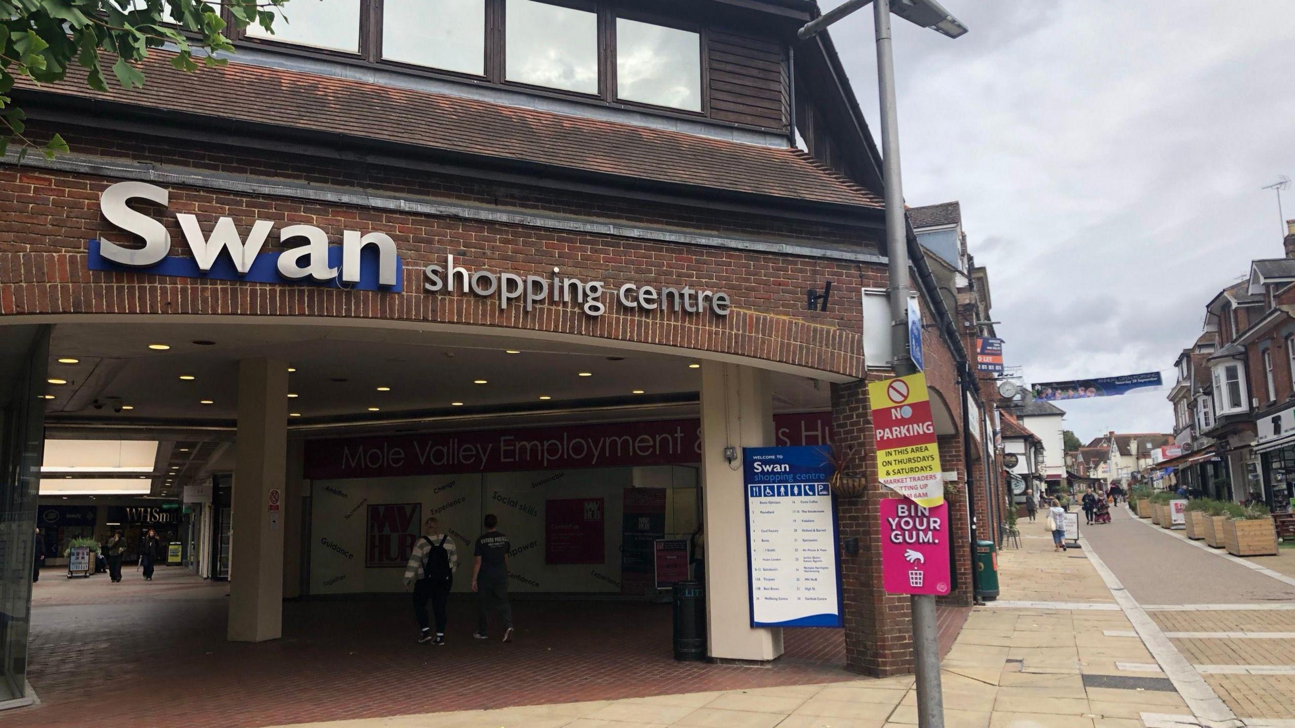 The entrance of a small brick shopping centre on a high street. The words Swan shopping centre are in silver lettering above an arched entrance