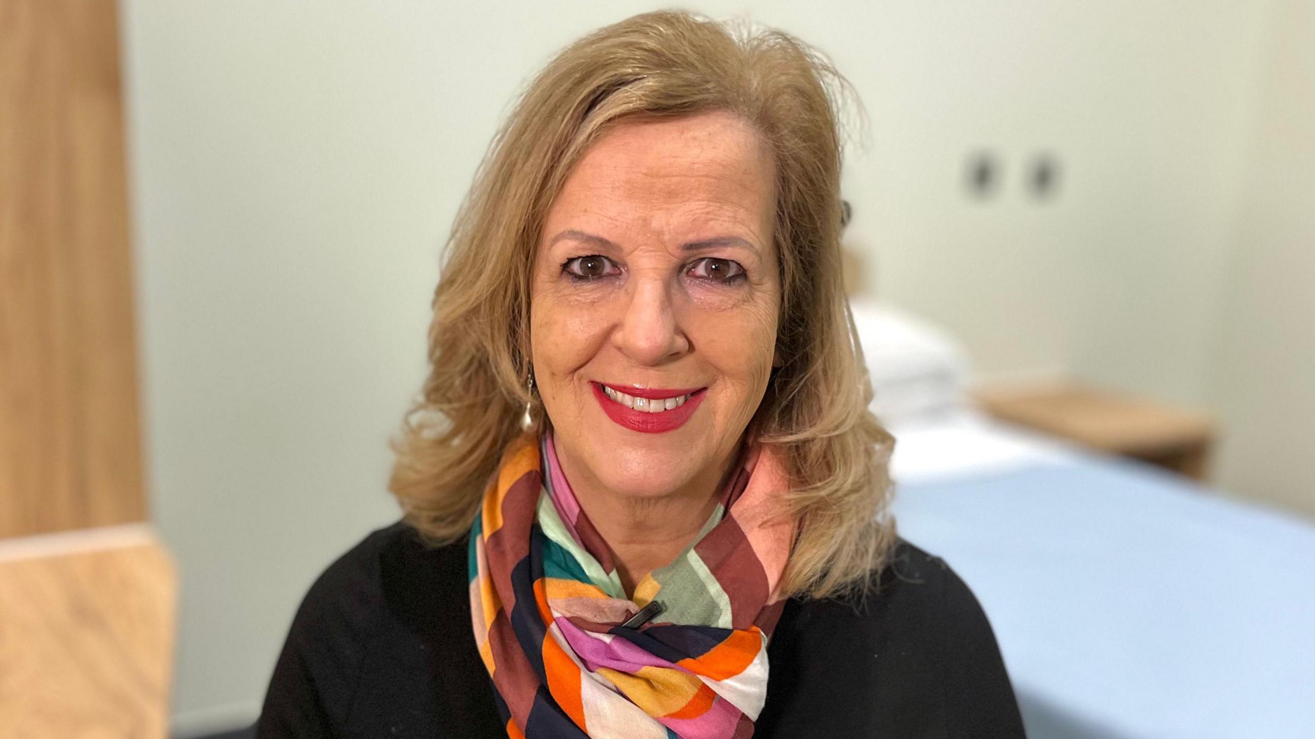 Caroline Dovan, a lady with blonde hair who is sitting down. She is looking directly at the camera and smiling and she is wearing a black T-shirt, with a coloured patterned scarf.