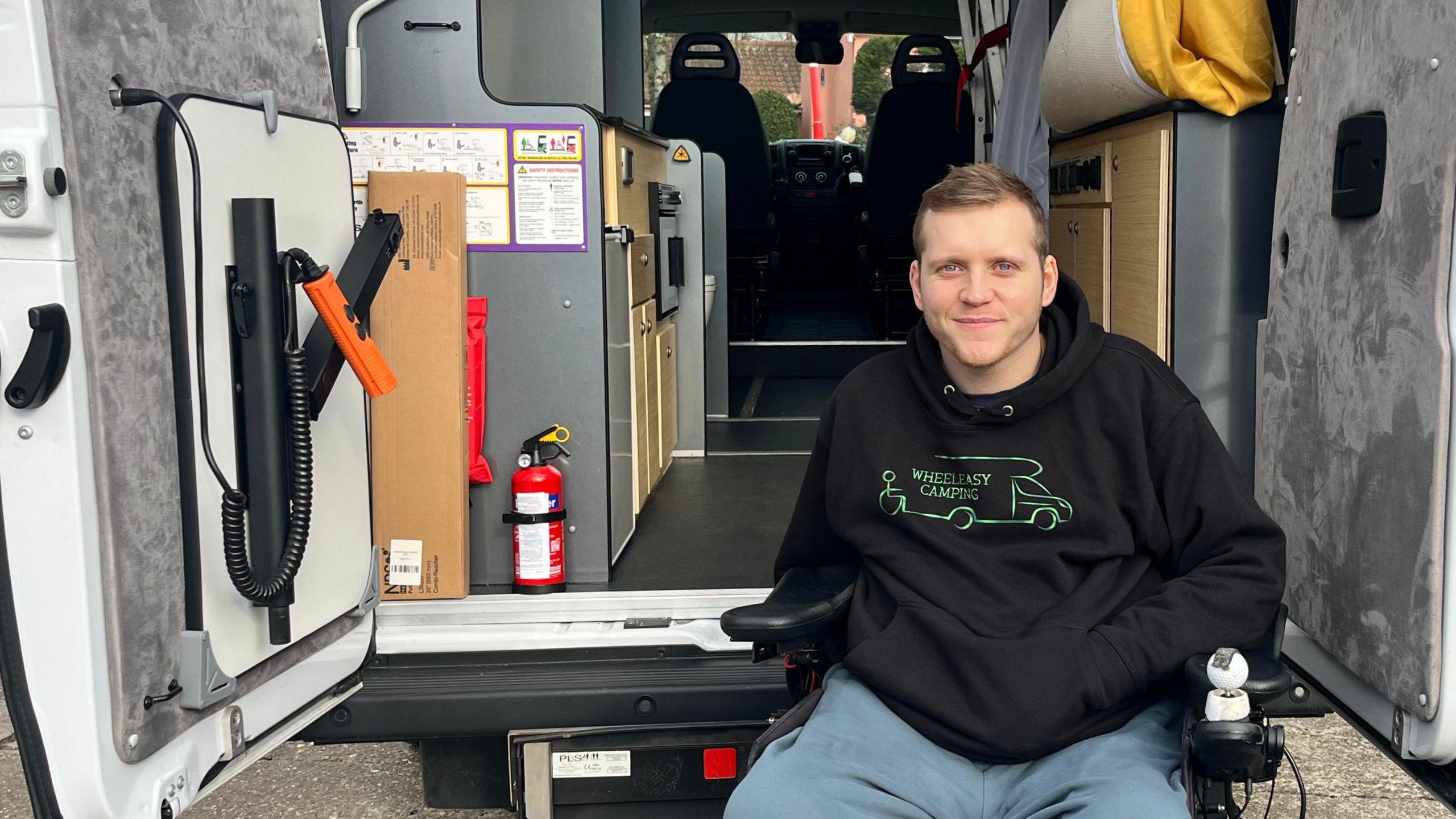 A young man with blonde hair and blue eyes, smiling, and sat in a black powerchair. He is wearing a black hoodie and blue trousers. Behind him, the back doors of his van are opened. At the back of the van, there are two seats. Around the van, there is concrete floors. On the right, there is a green hedge. 