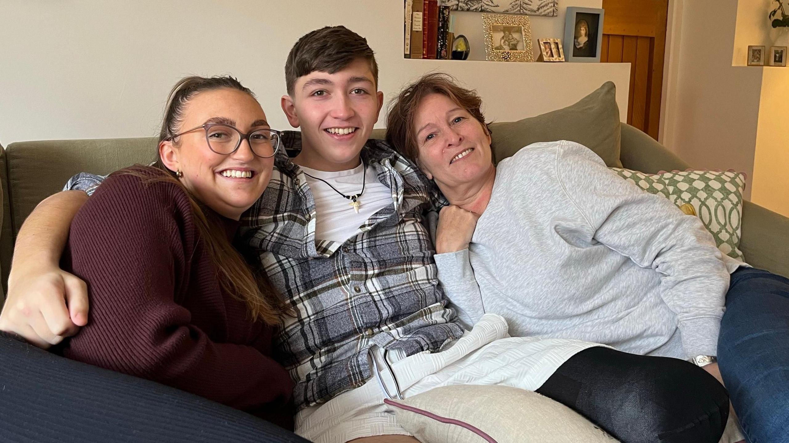 Ted with Jayme and his mother Sasha with his arms around them both sitting on the sofa in their living room. Jayme is wearing a maroon top, Ted is wearing a chequered shirt and has his amputated leg resting on a pillow with a black elastic sling on it. Sasha is wearing a grey jumper and all are smiling at the camera