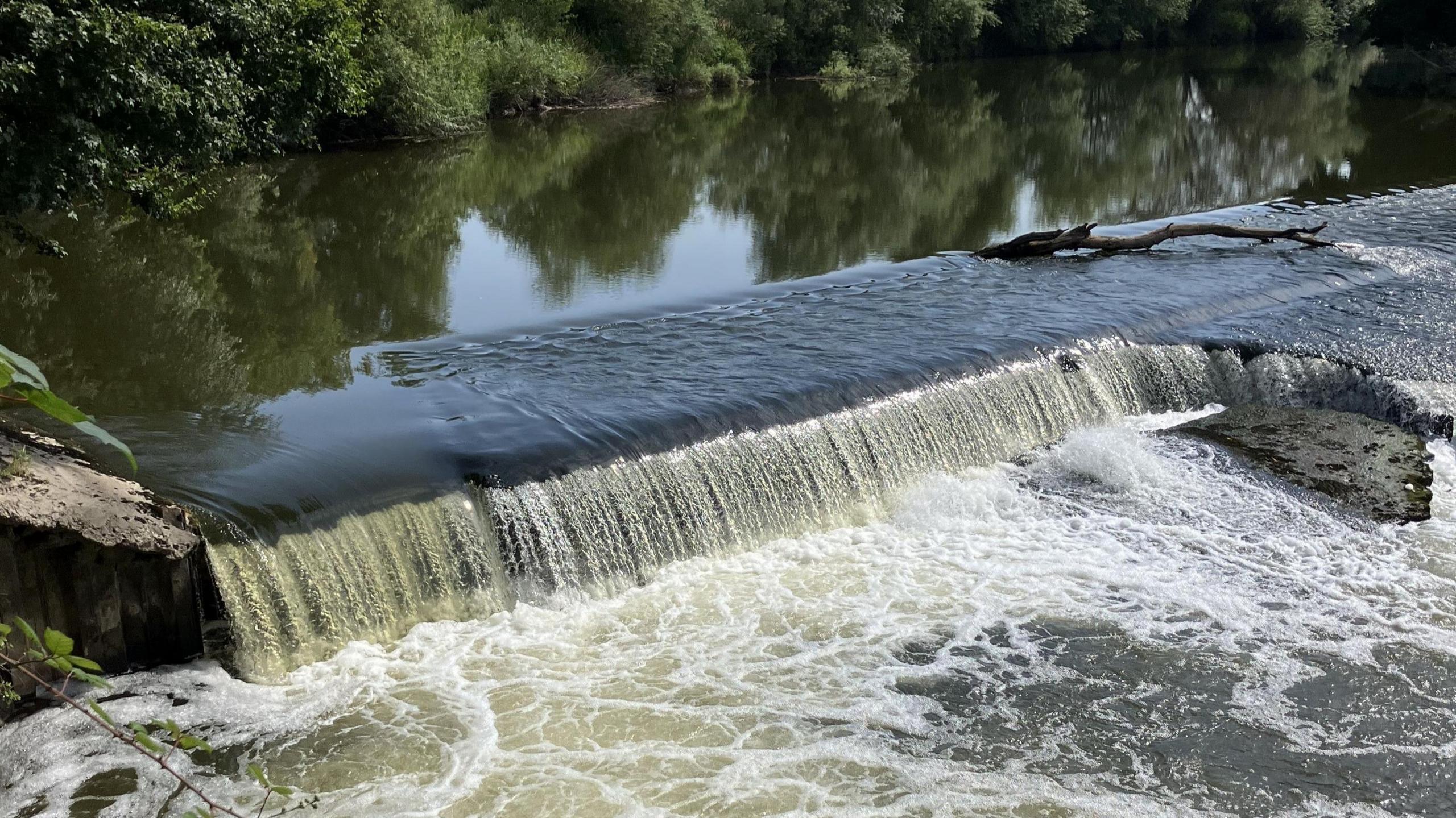 A photo showing a concrete weir spanning a river with water flowing over the top and green trees in the background