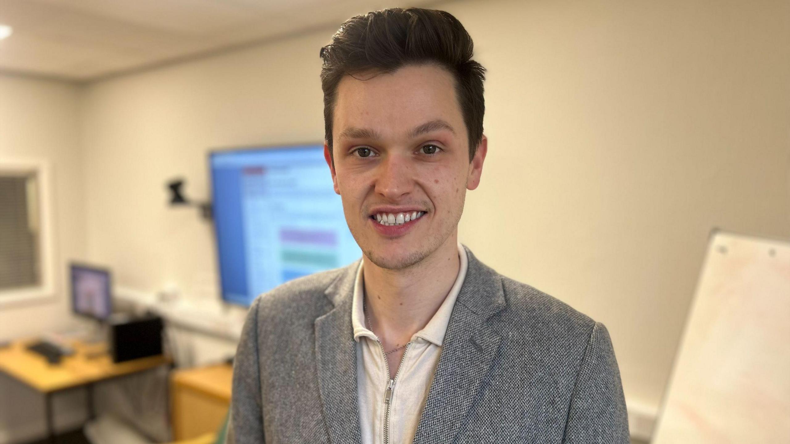 Ross Keat smiling. He is wearing a grey blazer and a creme zip-up top. He has short, dark hair. In the background a large TV and desk is blurred out.