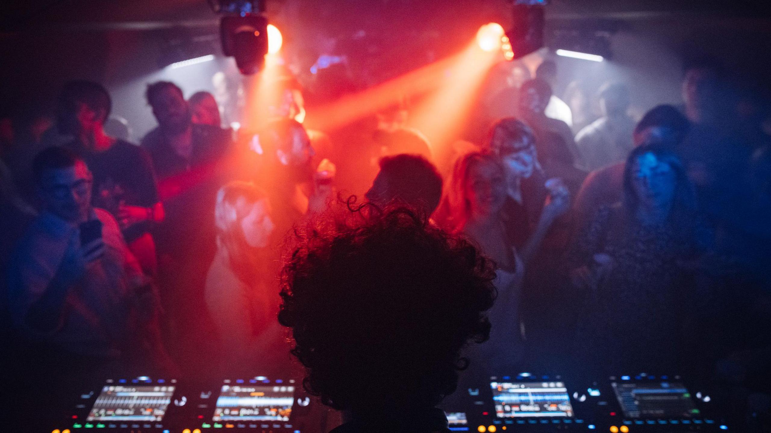 A DJ performs for the crowd in a gig at the Bristol Beacon cellars. They are viewed from behind lookout out to the crowd, who are lit up by spotlights