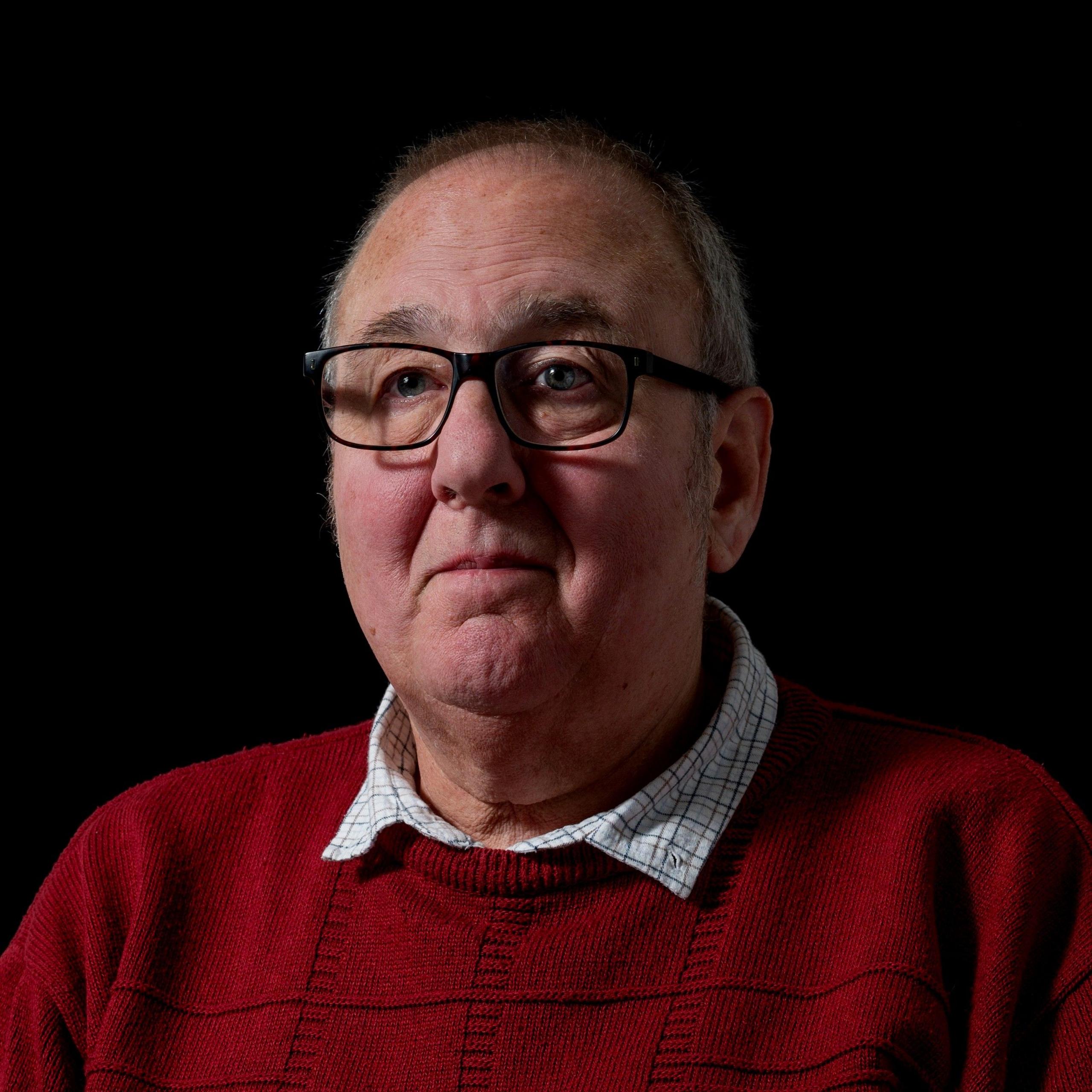 Michael Siddall, 66, portrait, looking slightly off left of camera, in front of a black background