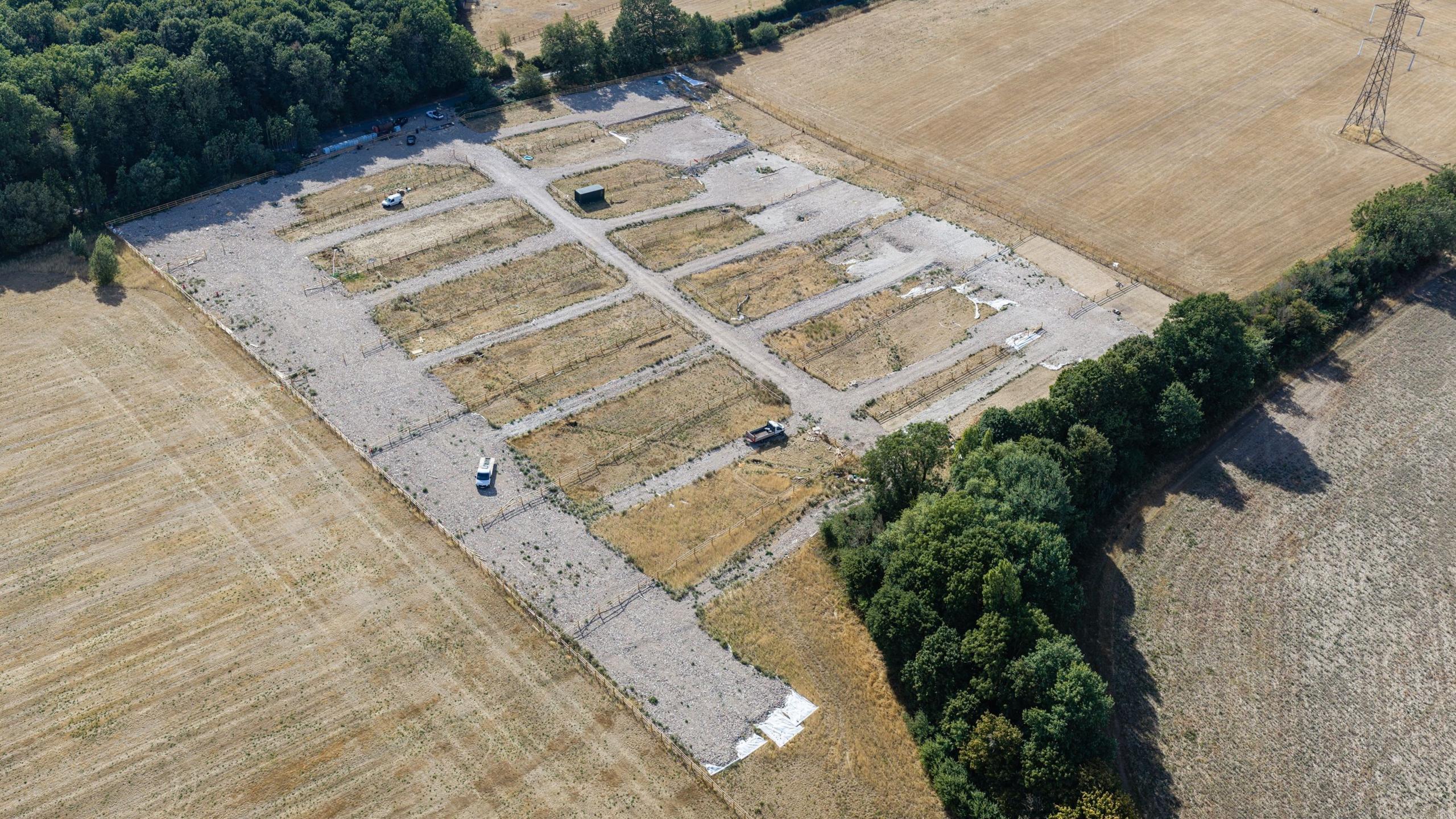 An aerial shot of the site, which is now clear of mobile homes and caravans, although two small vehicles can be seen. The hardstanding remains and the grass around it is now brown due to the drought. Green trees are at the edges.