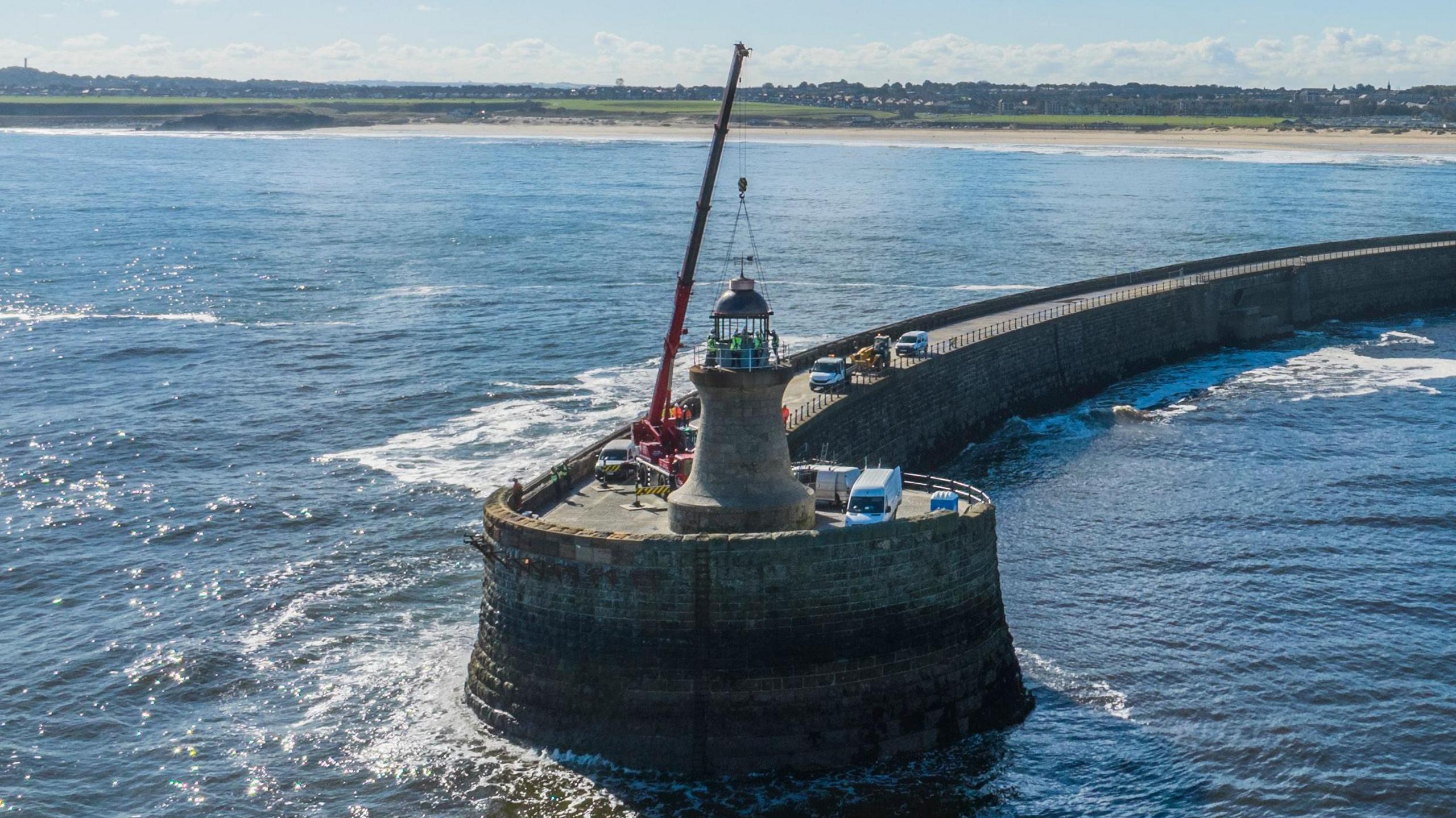 A red crane placing the new dome on relatively short, brick lighthouse. It sits on the circular end of a brick pier which arcs back to the shoreline. The lighthouse is surrounded by white vans and the coast is in the distance.