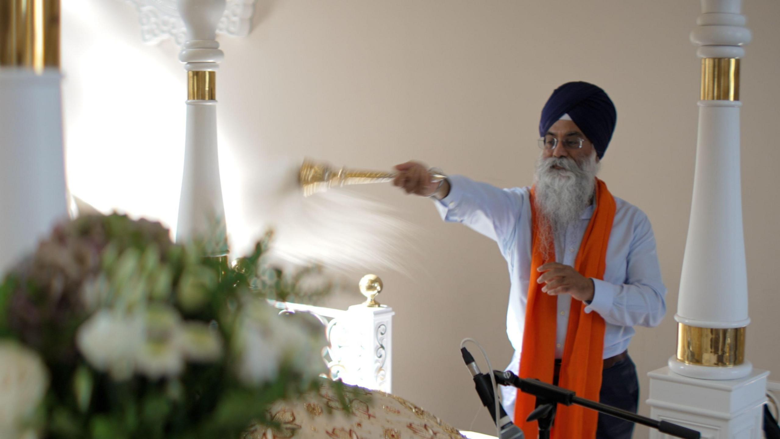 An older Sikh man stands in a temple. He is wearing a dark blue turban, a light blue shirt and a bright orange scarf-like garment around his neck. 