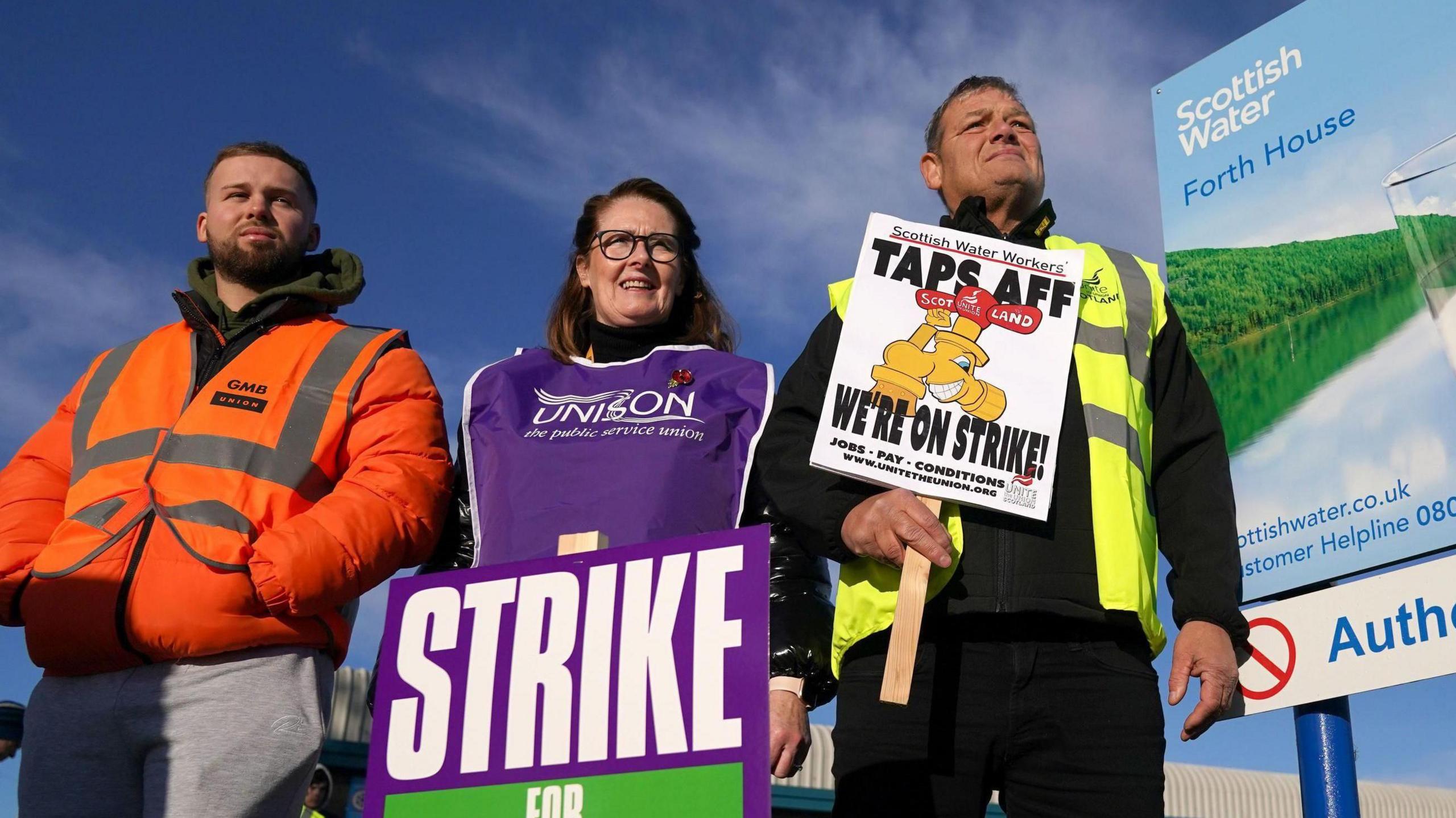 Two men and a woman stand on a picket line with a large Scottish Water billboard - featuring a glass of water against a scenic backdrop - to their right.