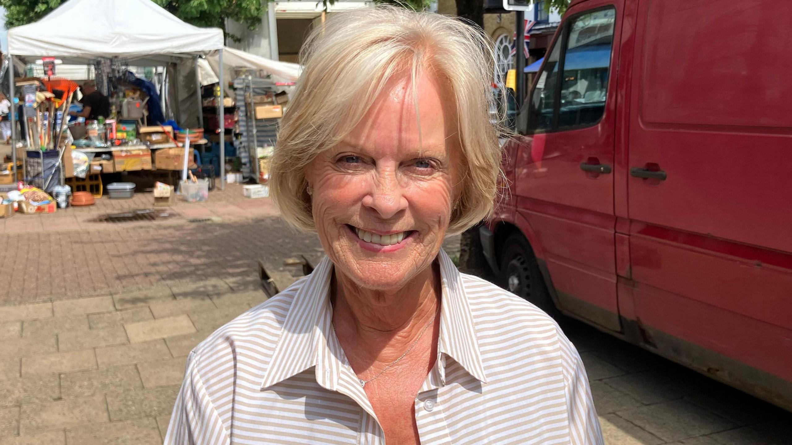Pauline Ashton is smiling at the camera in a market square with stalls and a van behind her. She has a blonde bob and is wearing a grey-striped blouse