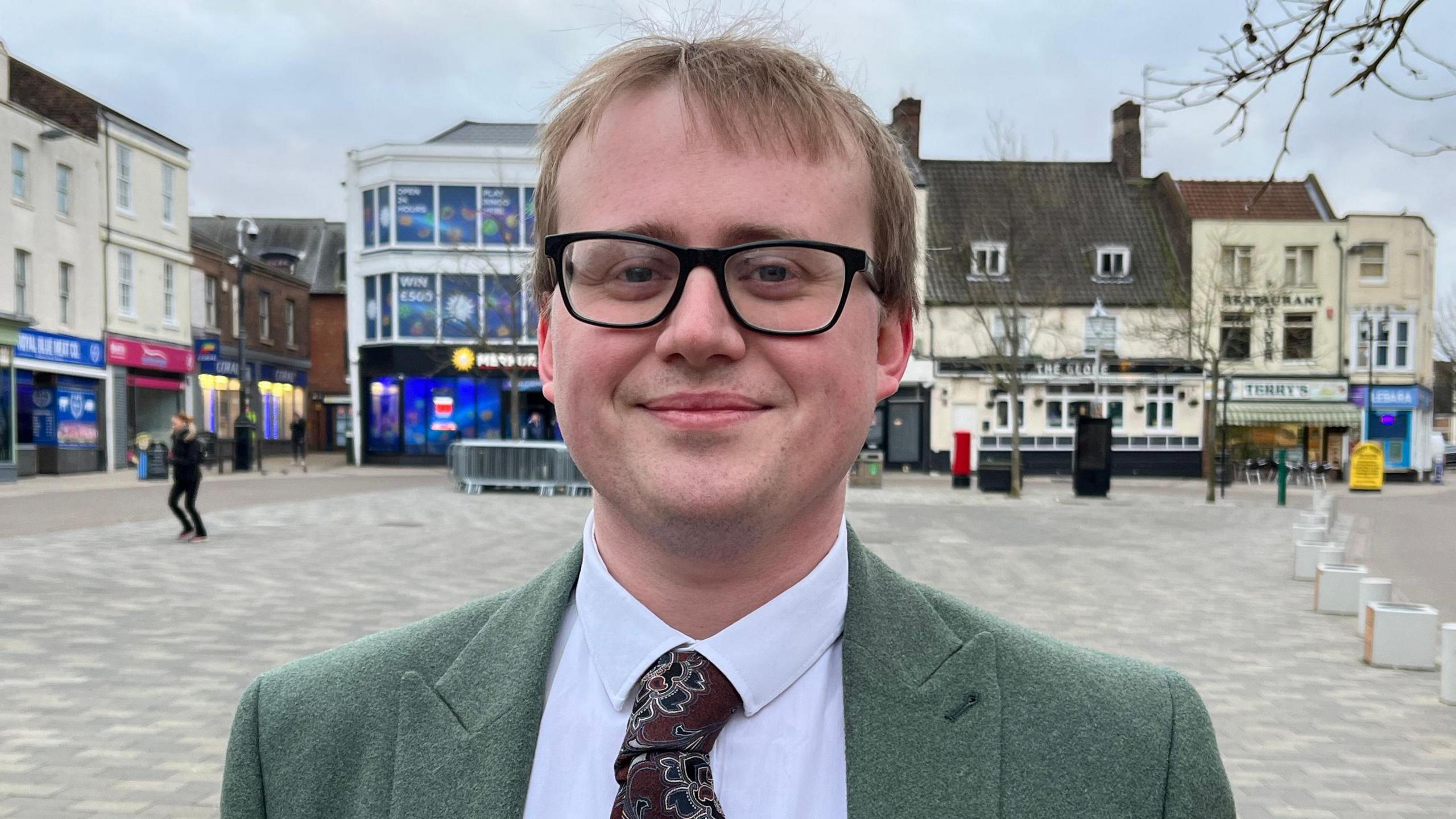 Feed Fenland founder George Broughton wearing a green suit jack with a paisley tie smiles at the camera as he stand in the middle of Wisbech Market Square.