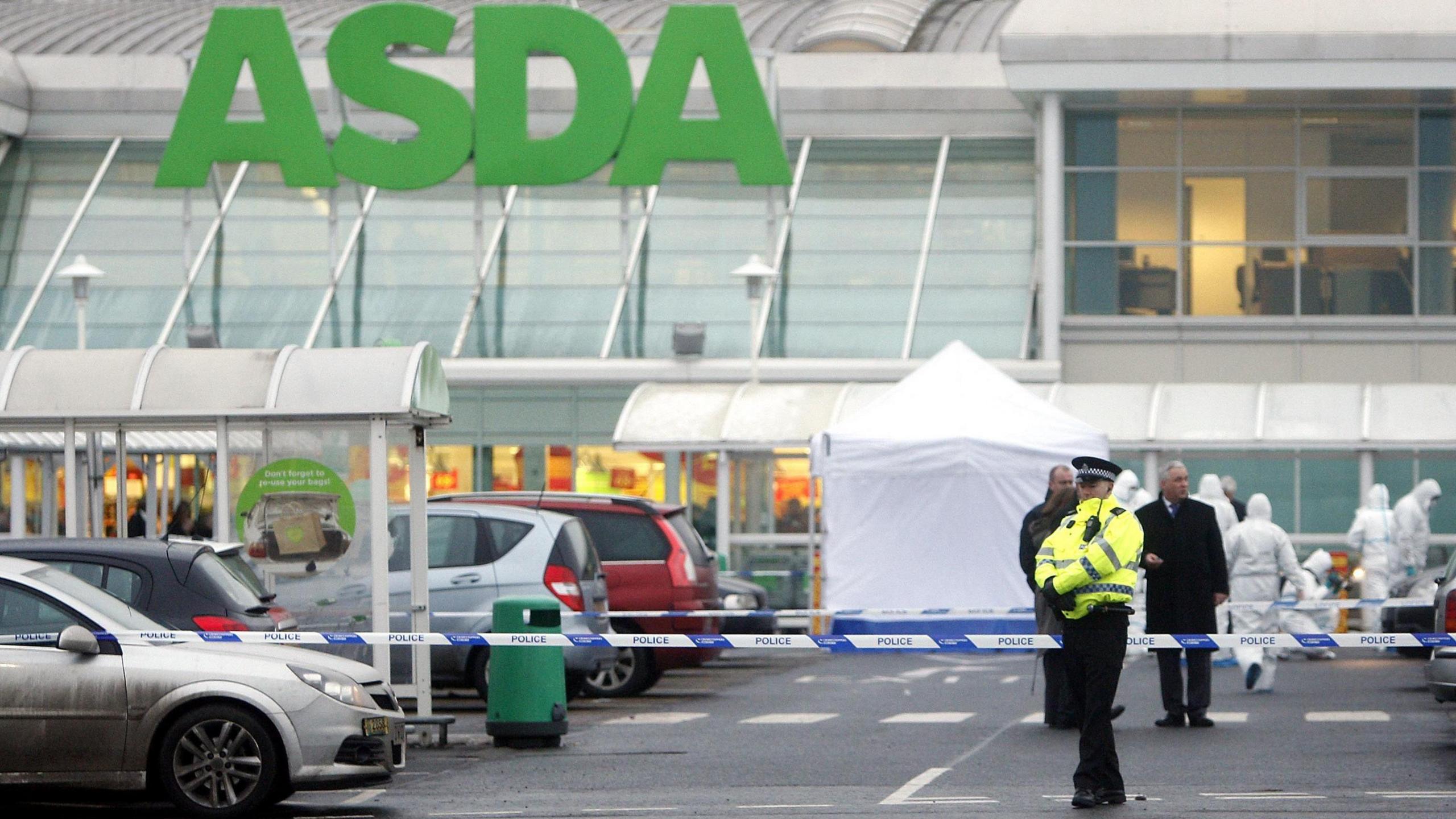 A lone police officer stands guard in front of blue and white police tape in the car park of Asda, Robroyston. Behind the tape a white forensic tent has been erected and specialists in white hazmat suits can be seen carrying out inquiries. Plain clothes detectives, including one wearing black trousers and a long black coat, are also visible in the background.