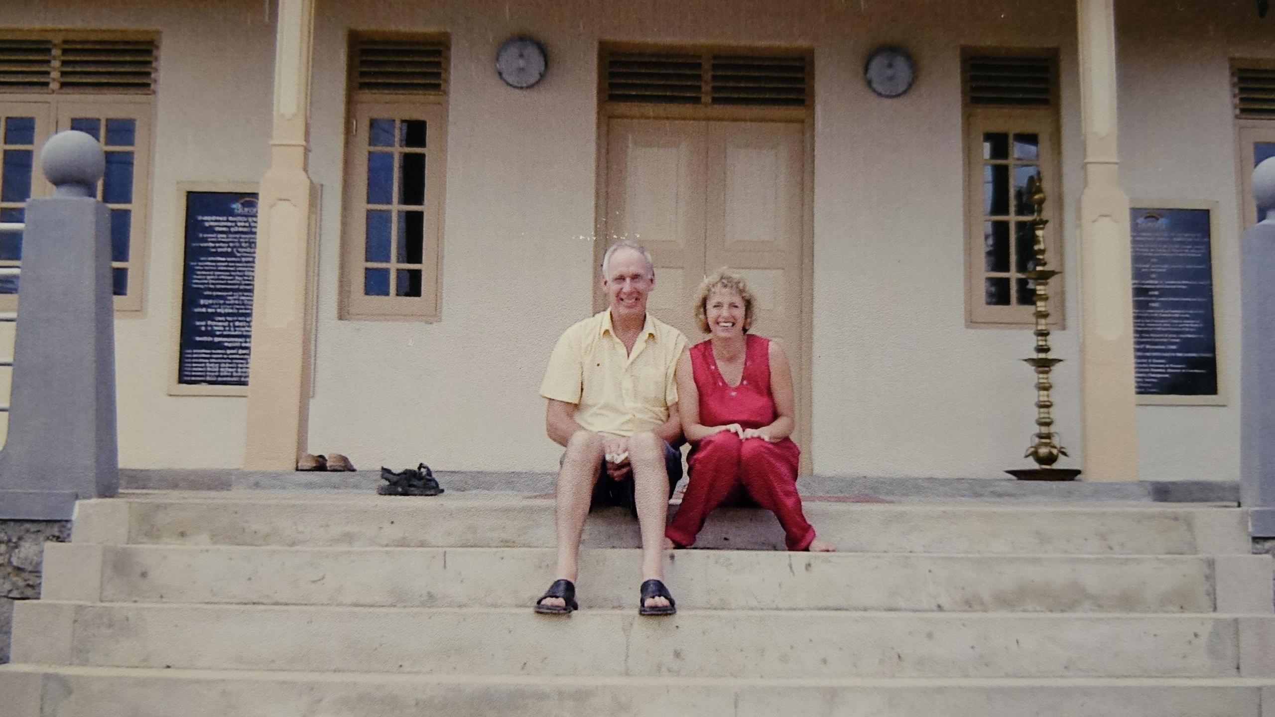 A man in a shirt and shorts and a woman in a red top and trousers sit on the stairs in front of a yellow building.