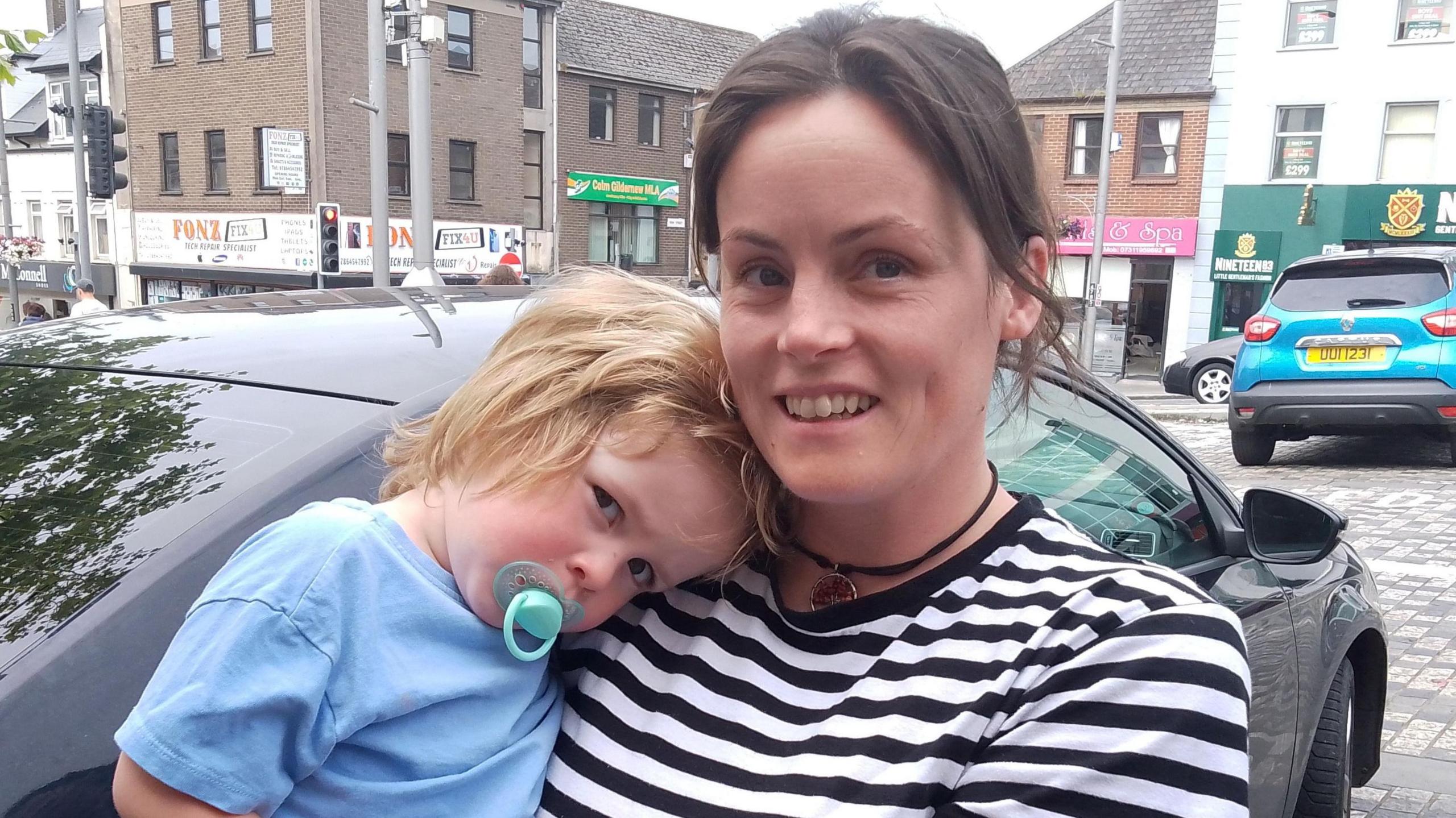 Lynn is holding her son Rian.  She is wearing a black and white stripped t-shirt. Rian is wearing a blue top and has a green dummy in his mouth. They're standing on a street with cars behind them.