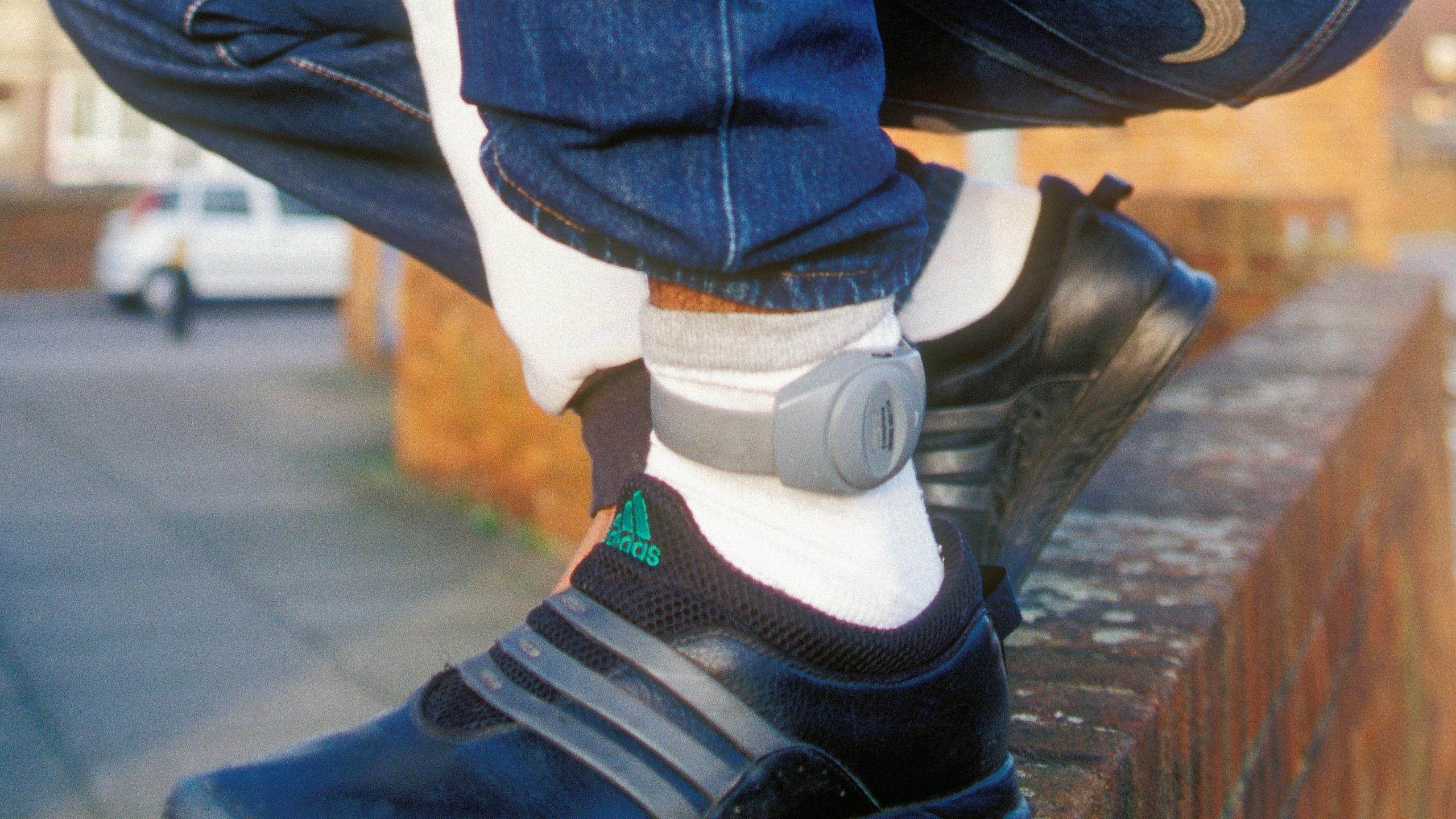 A grey, electronic tag being worrn round the ankle of someone wearing white socks and black trainers, perched on a wall.