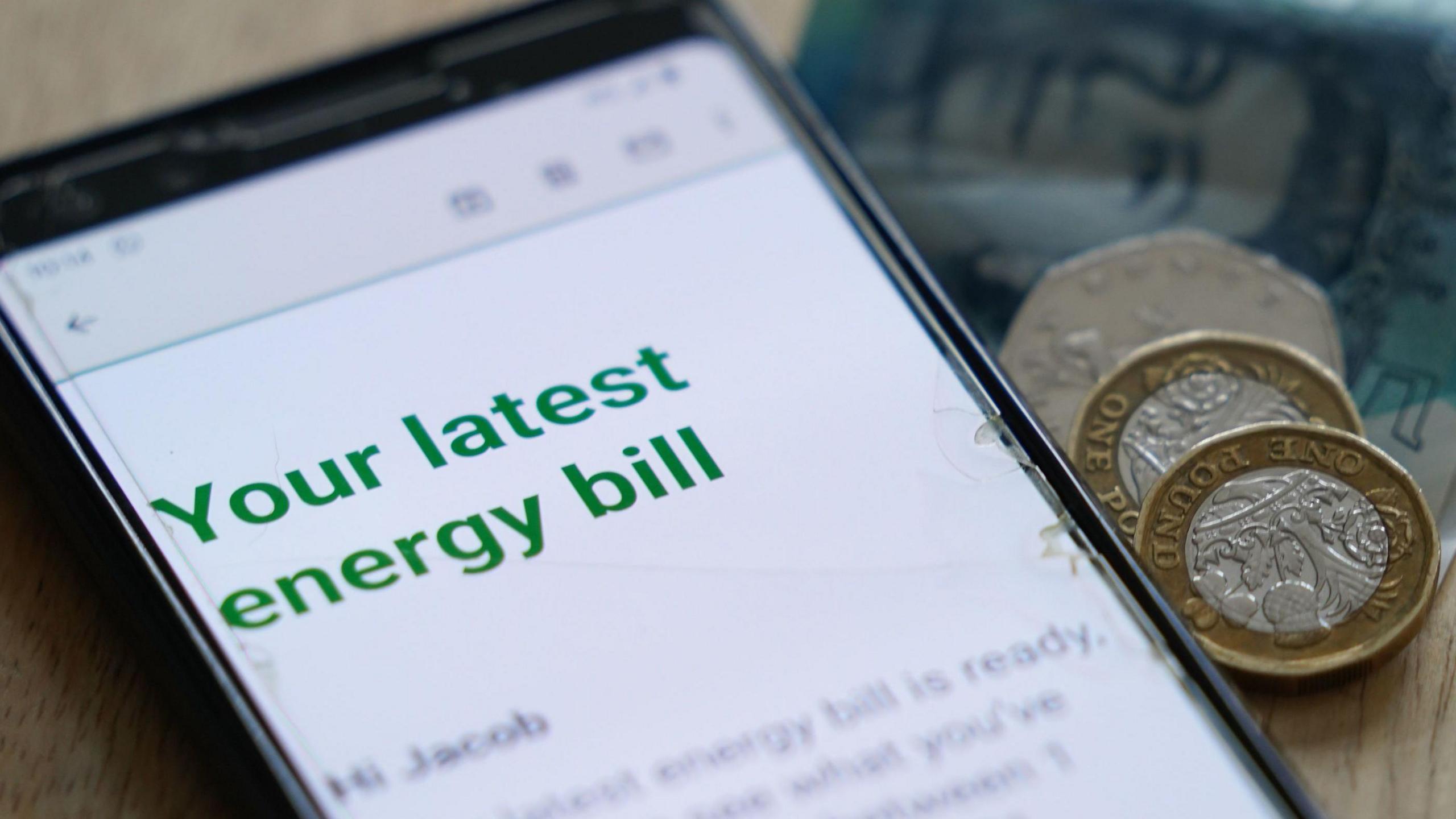 A close up of a mobile phone with the message "Your Latest Energy Bill". Beside the phone are two pound coins and a 50p coin.