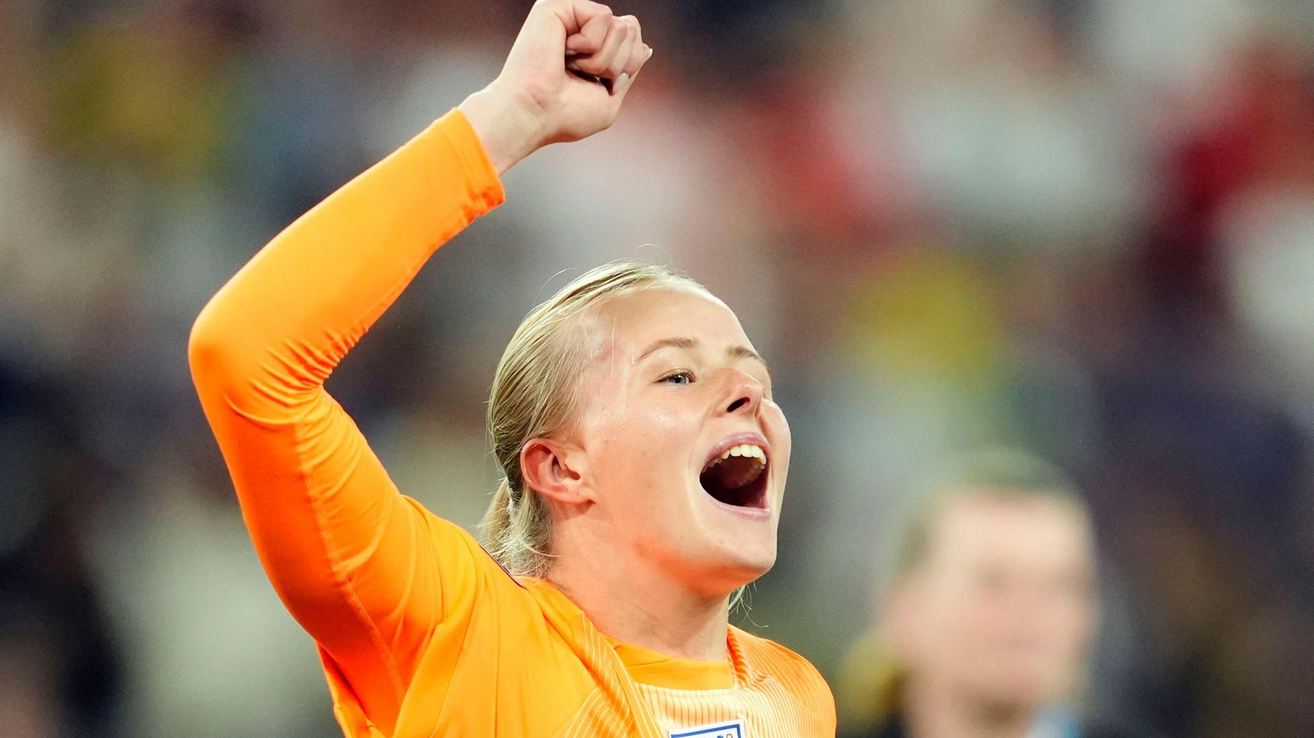 A woman in an orange goalkeeper kit celebrates with one arm in air on the pitch following a match. She has blonde hair tied back and her mouth is open mid-shout.
