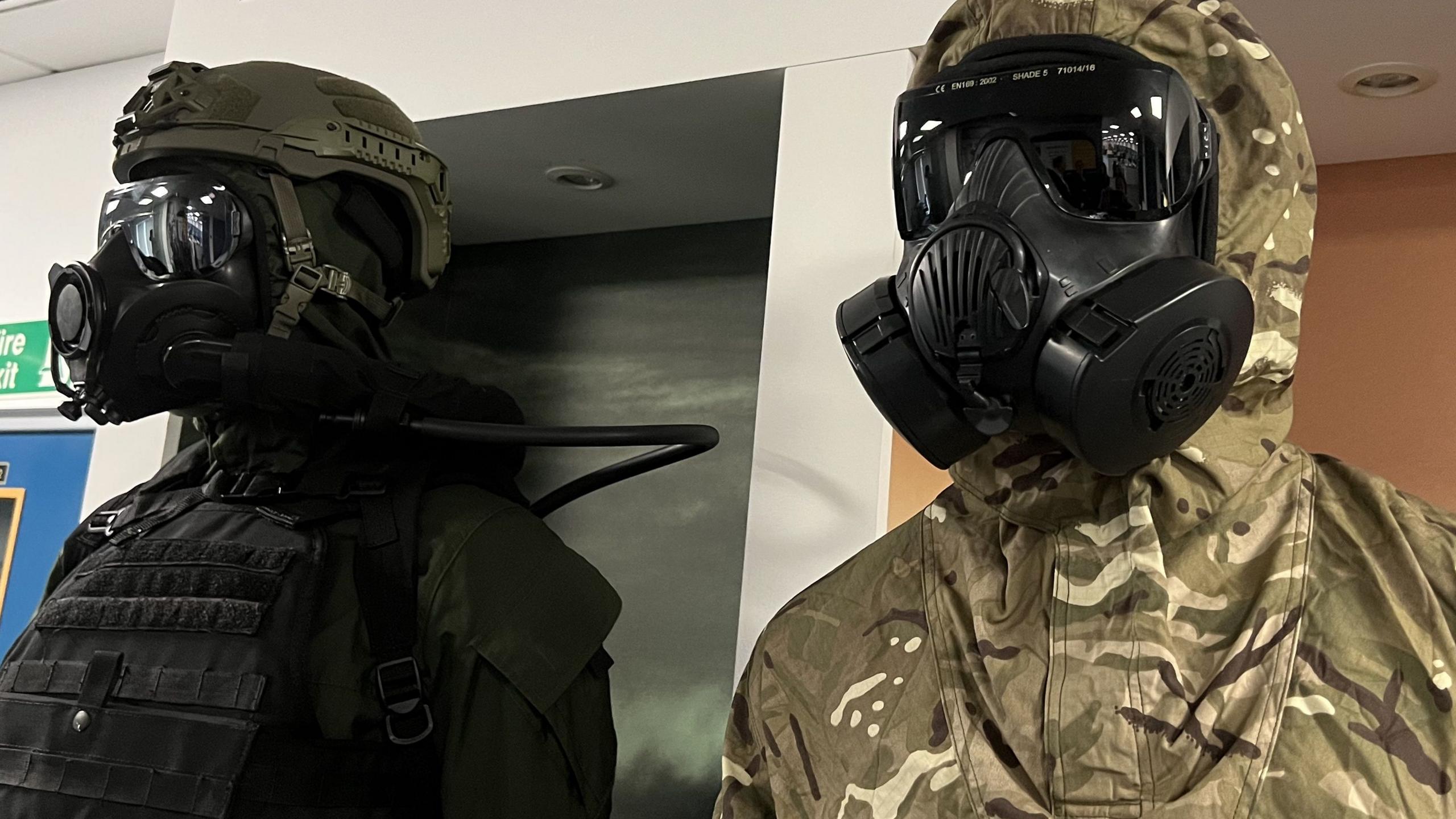 Two mannequins standing side by side. One is in military protective equipment the other is in black special forces equipment. Both have black, modern gas masks covering their entire faces. 