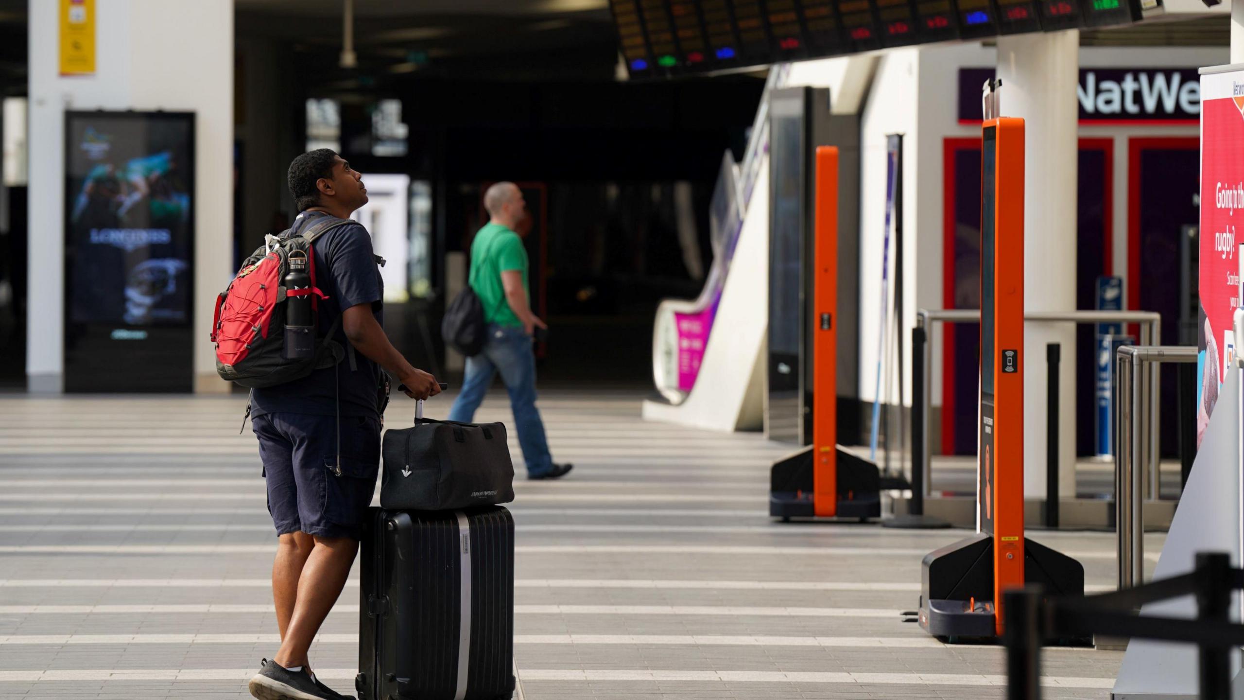A passenger looks at message boards at Birmingham New Street Station during strike action in 2022. The man has a case and smaller bag and is carrying a rucksack. Behind him another man wearing a green t-shirt and jeans is walking along. 