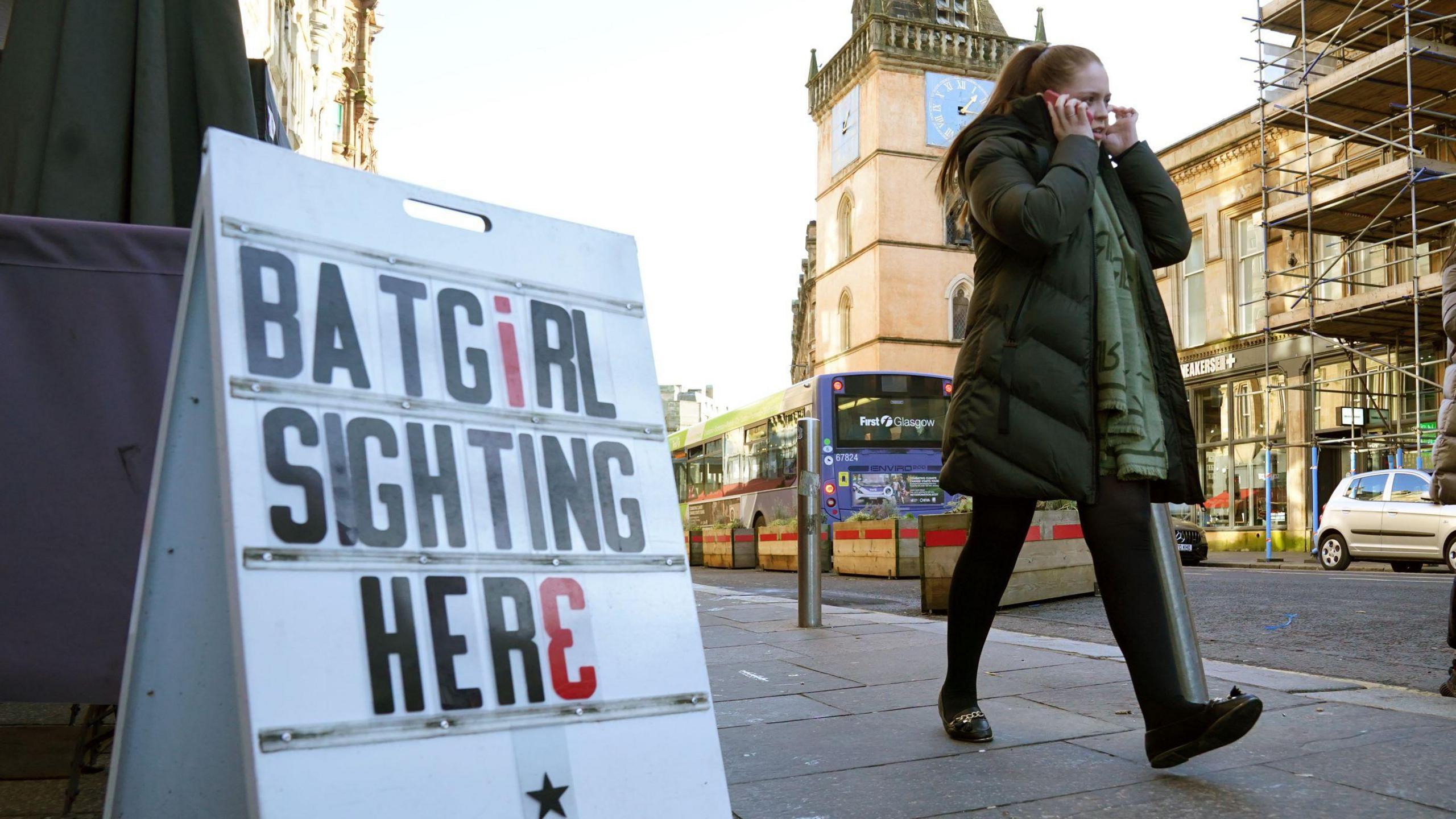 A woman walks past a prop sign from the Batgirl film, which reads BATGIRL SIGHTING HERE. In the background traffic is going past.