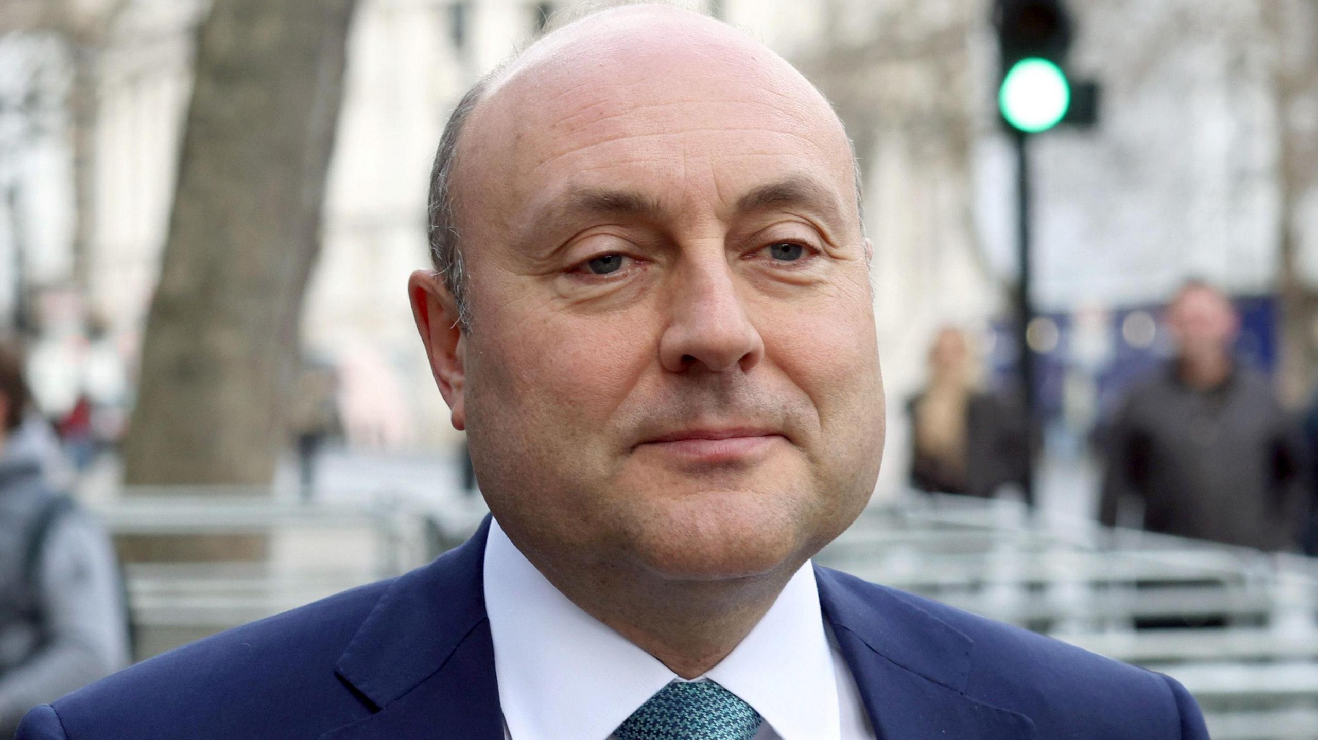 A man is standing in front of the camera but nt looking into the lense. He's wearing a navy suit with a white shirt and a tie. The background is blurred.