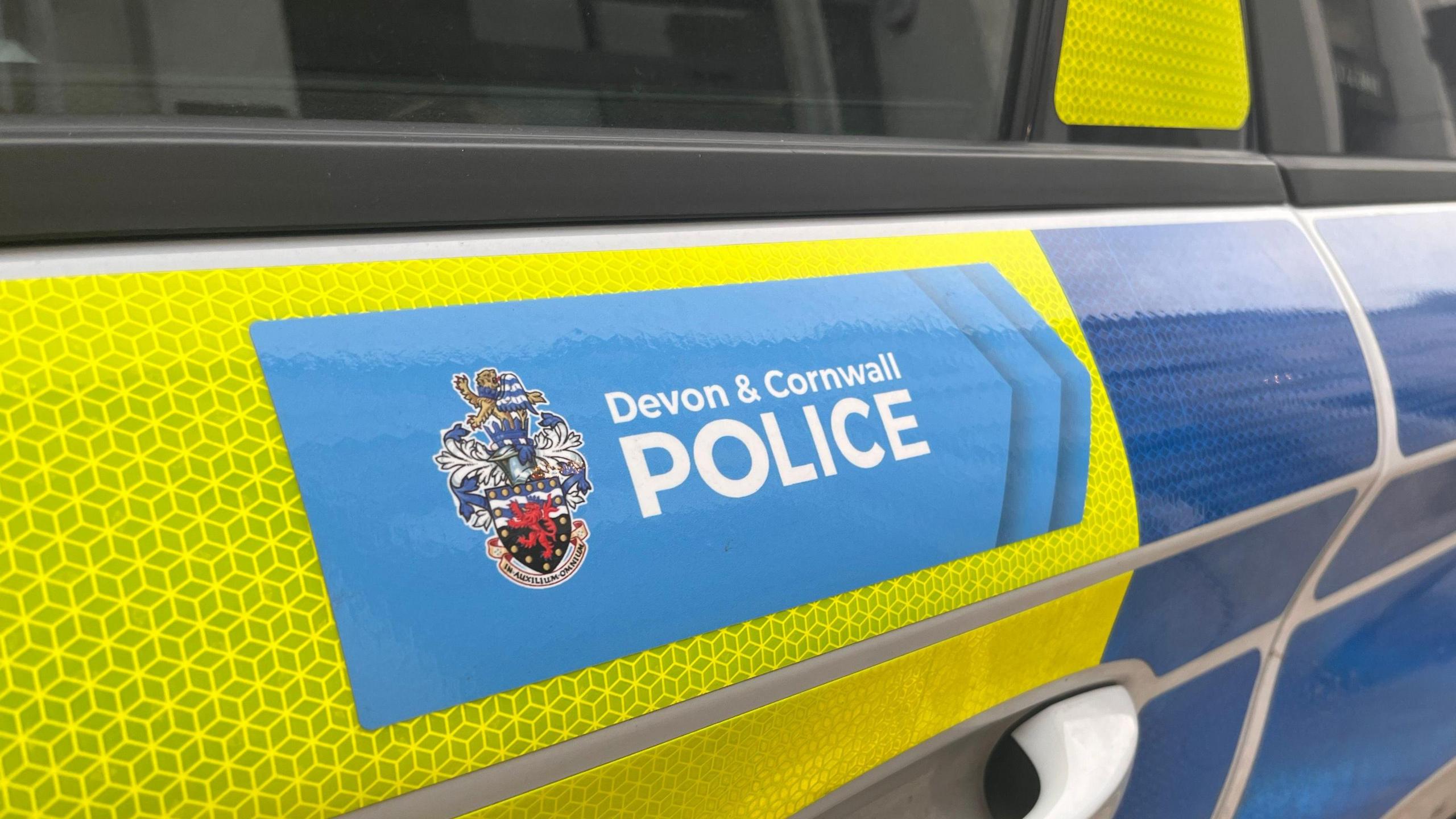 The door of a police car with reflective yellow and blue strips on it and a Devon and Cornwall Police logo.