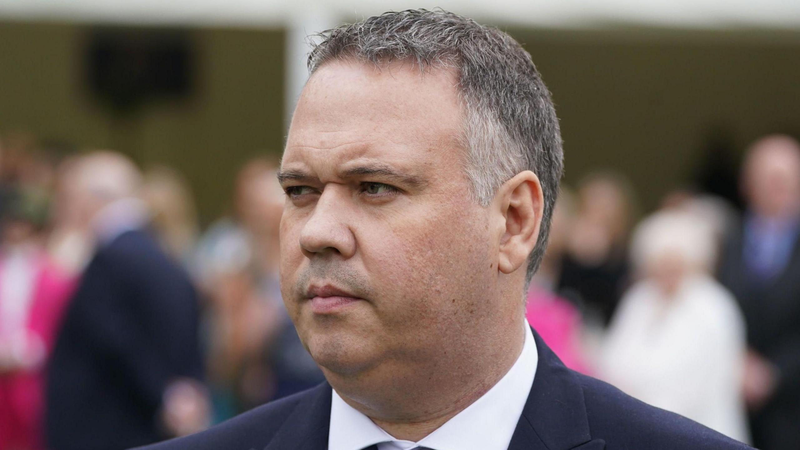 A close up image of John Caldwell, wearing a white shirt and navy suit. He has short grey hair. The background of the image is blurry.