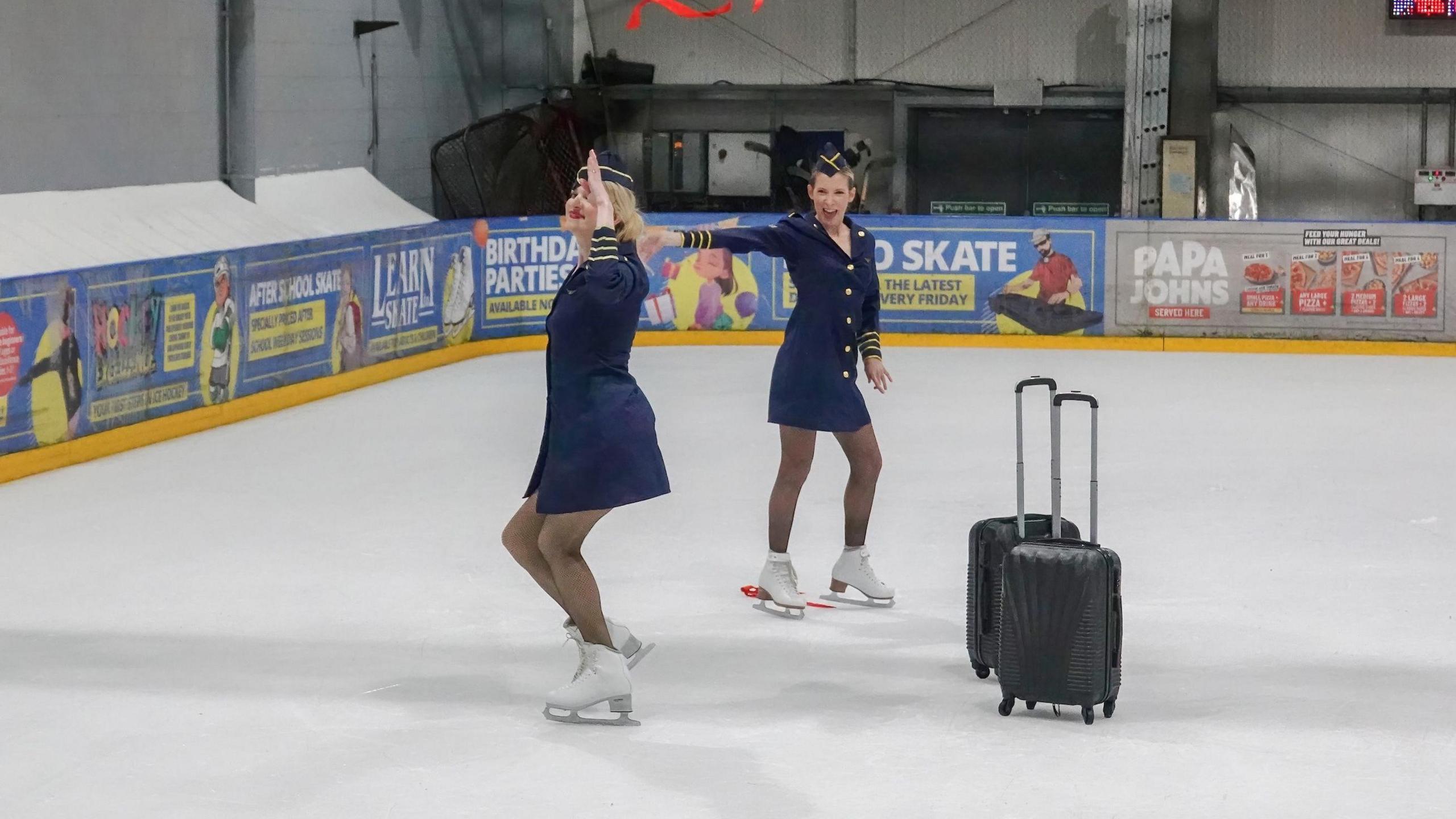 Two women dressed in blue air hostess uniforms and ice skates stand on an ice rink while performing. There are two black suitcases next to them on the rink.