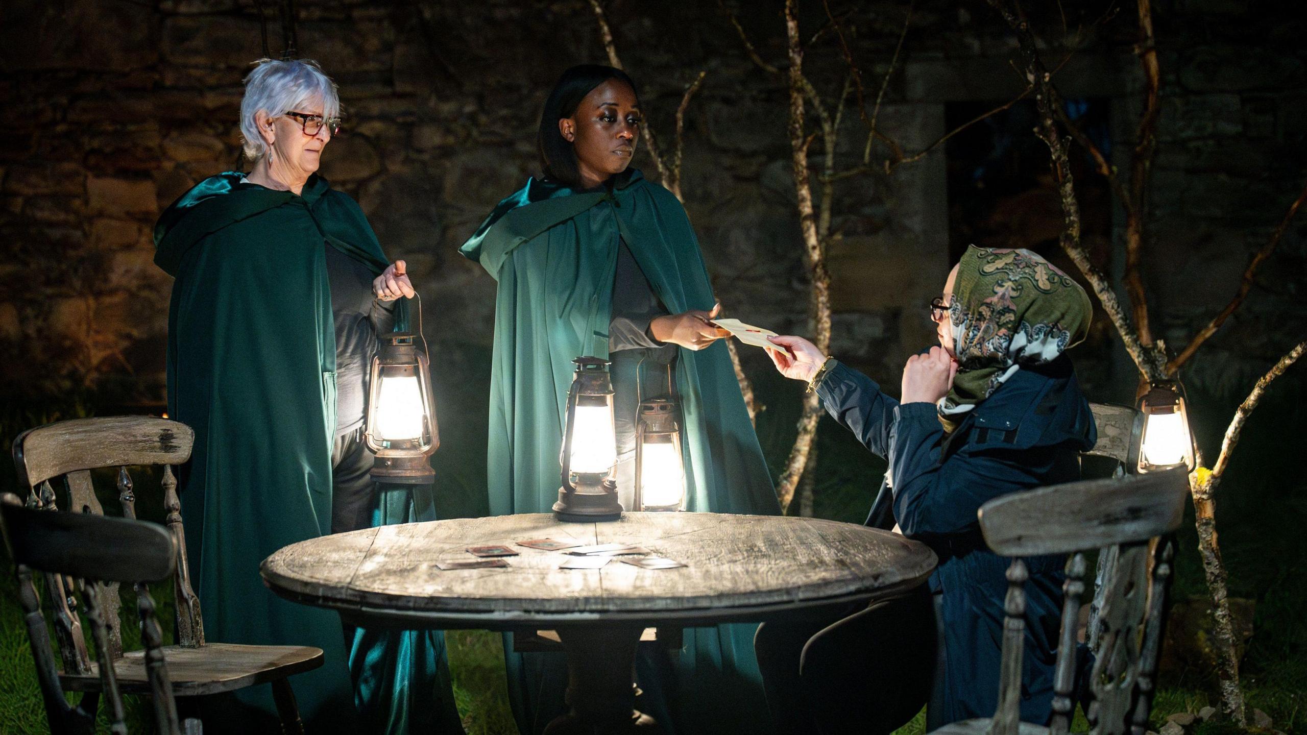 Linda and Minah dressed in long green cloaks handing a rolled up piece of paper to Fozia at a torch-lit wooden table outside.