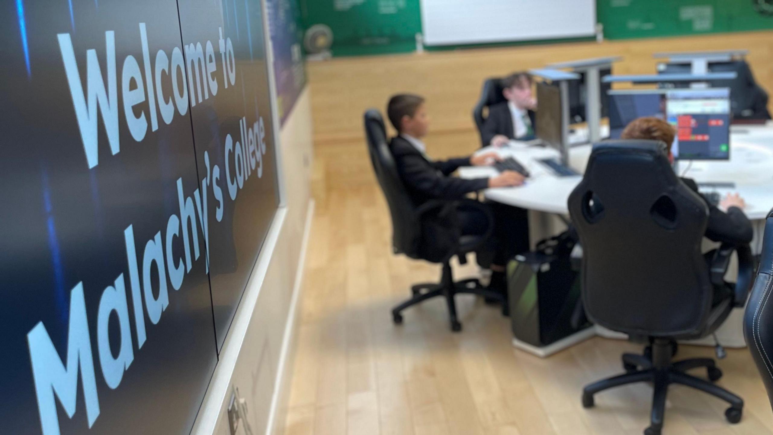 Photo of a school classroom with students sat at grey circular desk with multiple computer screens. 

Focused on the image is a black welcome display board at the side of the classroom. 