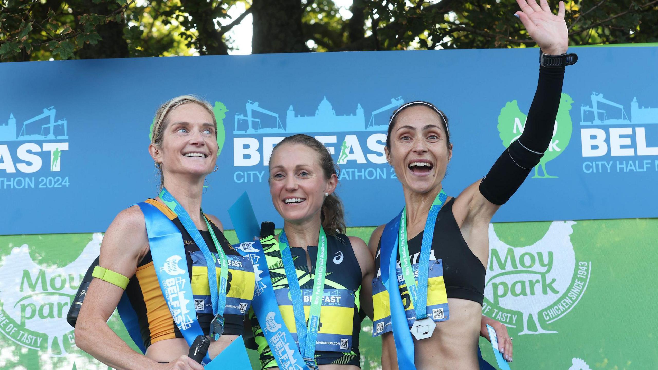 Three women smiling and cheering on the podium after the race, each with their medals. 