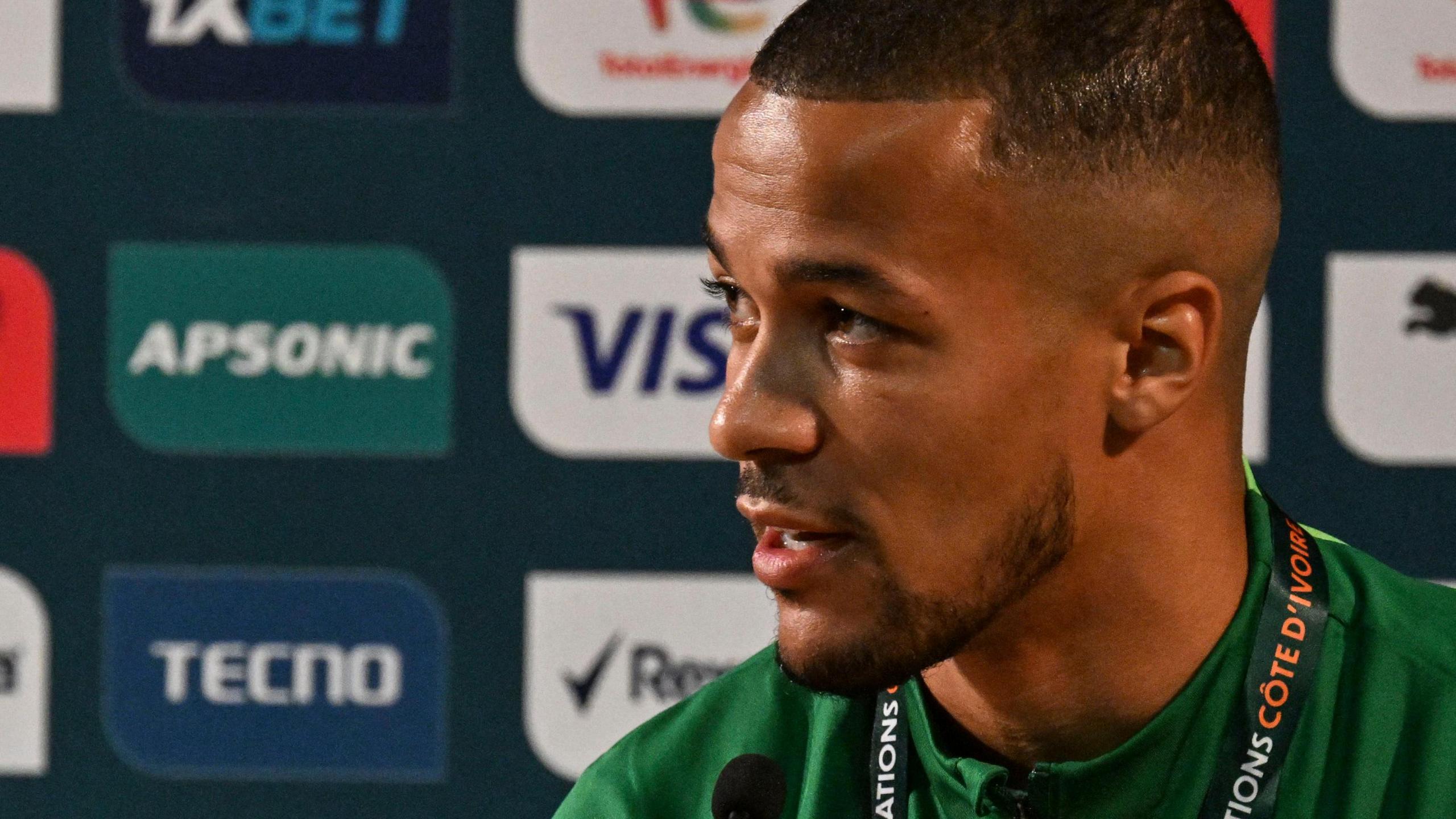 A headshot of William Troost-Ekong looking to his right, wearing a green top and with a lanyard around his neck, talking in front of a sponsors' advertising board that has various logos on it.