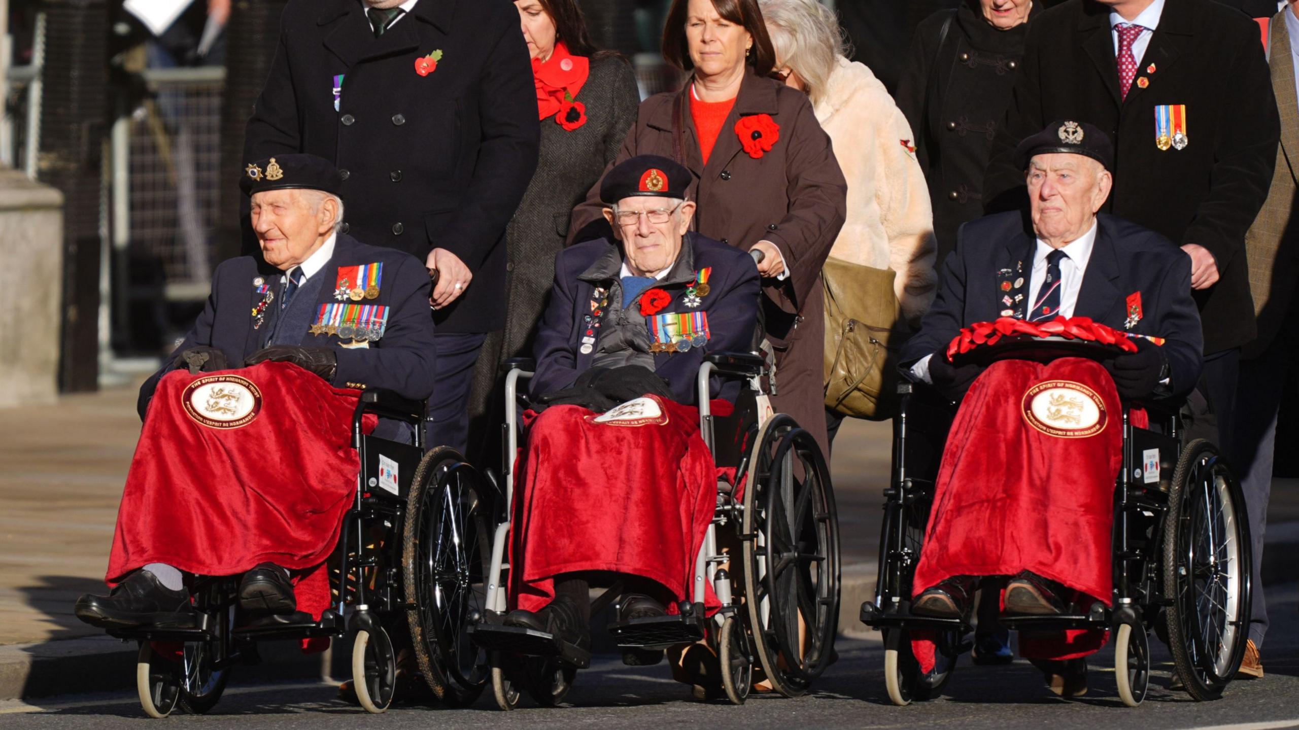 Three World War Two veterans in wheelchairs, with red blankets over their legs, being pushed by relatives wearing poppies in central London